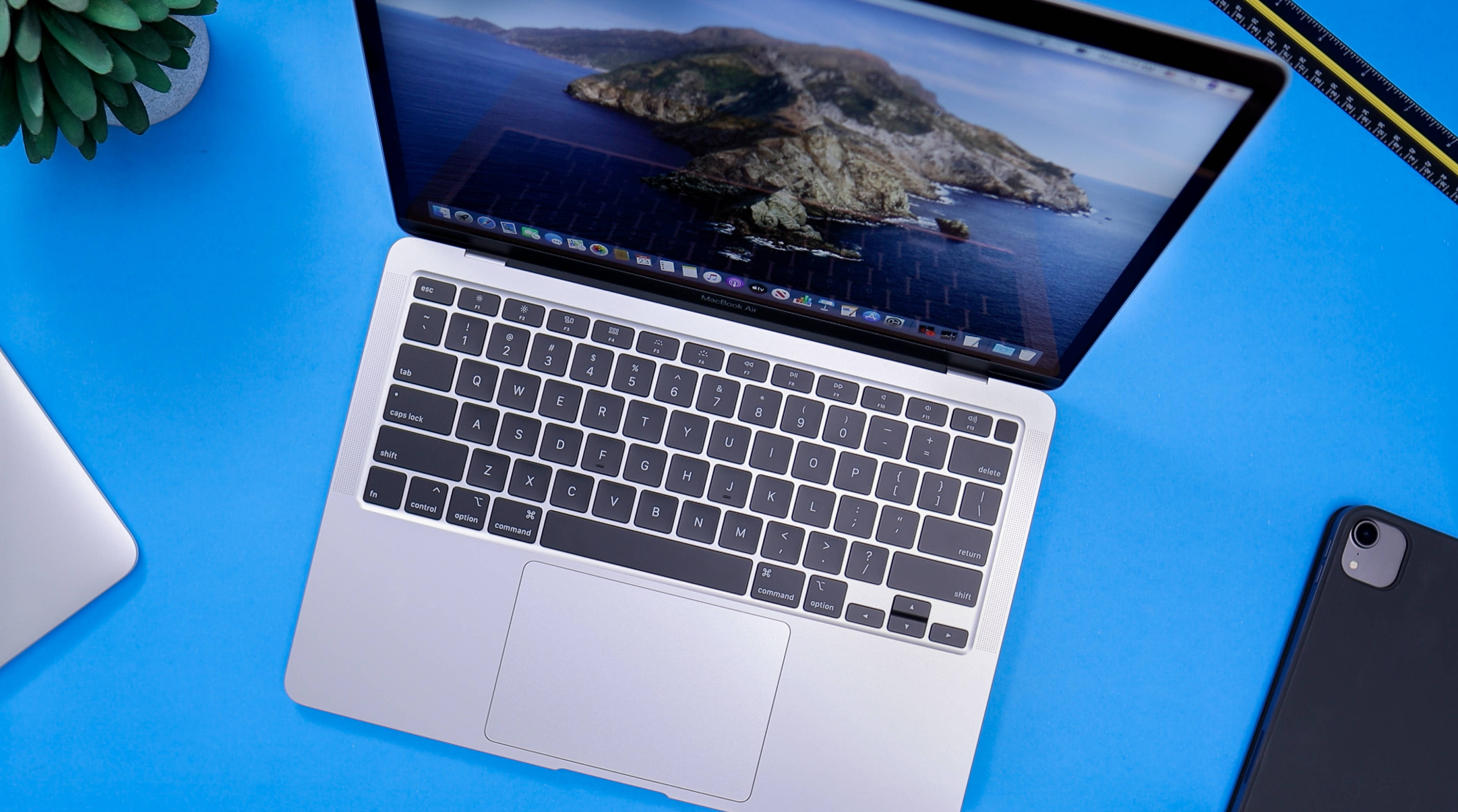 Macbook Air 2020 桌布 壁紙 — 銀色筆記型電腦放在亮藍色書桌上，附近有島嶼壁紙和多肉植物。