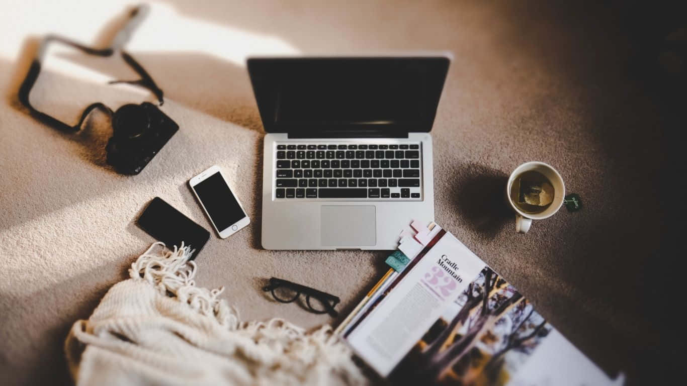 Laptop 1366 X 768 桌布 壁紙 — 鋪有地毯的地板上有一個舒適的工作空間，配有筆記型電腦、電話、眼鏡和一杯冒著熱氣的茶，沐浴在溫暖的陽光下。