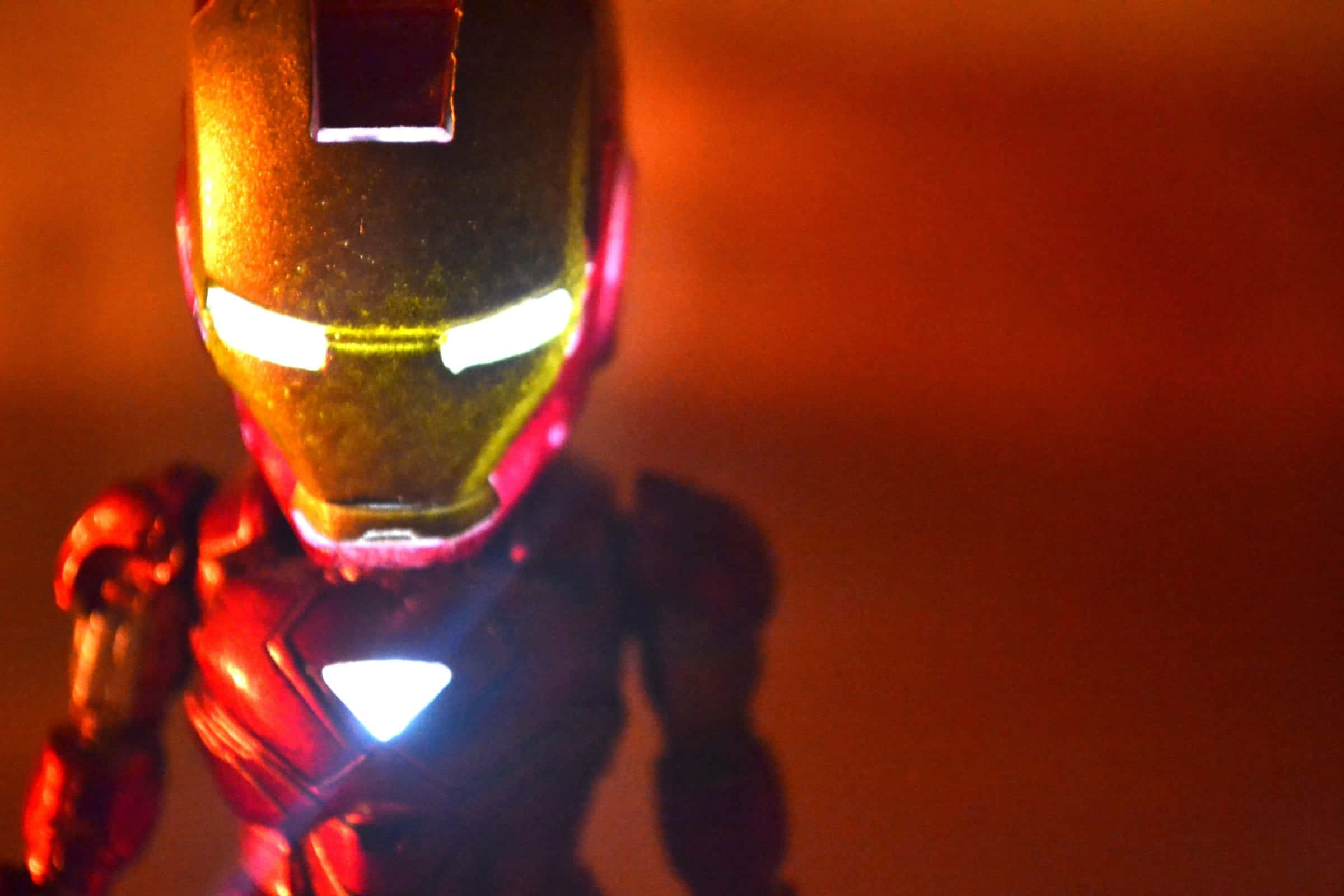 Iron Man Action Figures 桌布 壁紙 — 鋼鐵人動作人偶的特寫，眼睛和胸部弧形反應器發光，背景為溫暖的橙色。