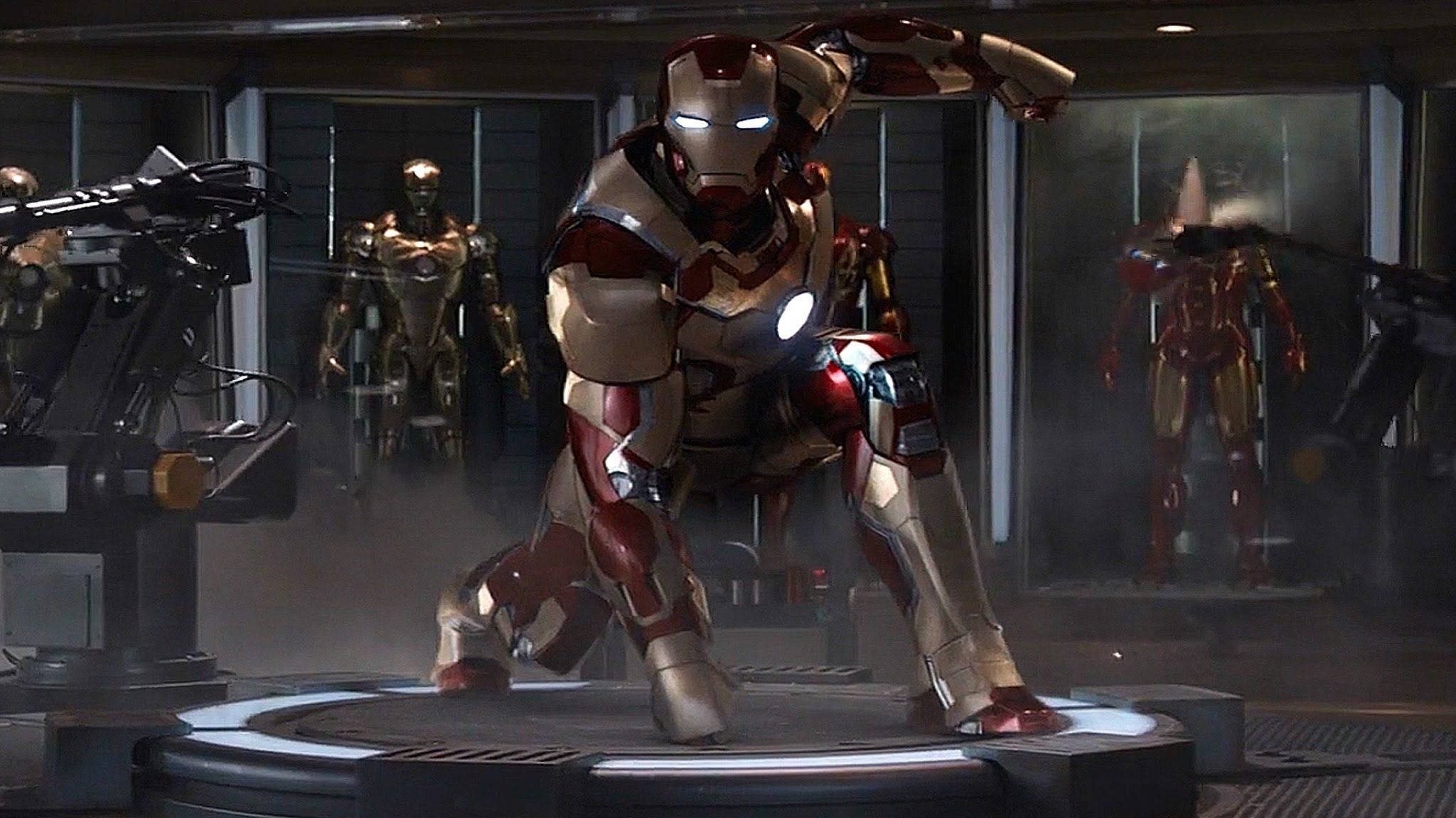 Iron Man Mark 3 桌布 壁紙 — 鋼鐵人穿著紅金色裝甲，蹲在一個高科技實驗室的圓形平台上，身後展示櫃中還有其他戰衣。