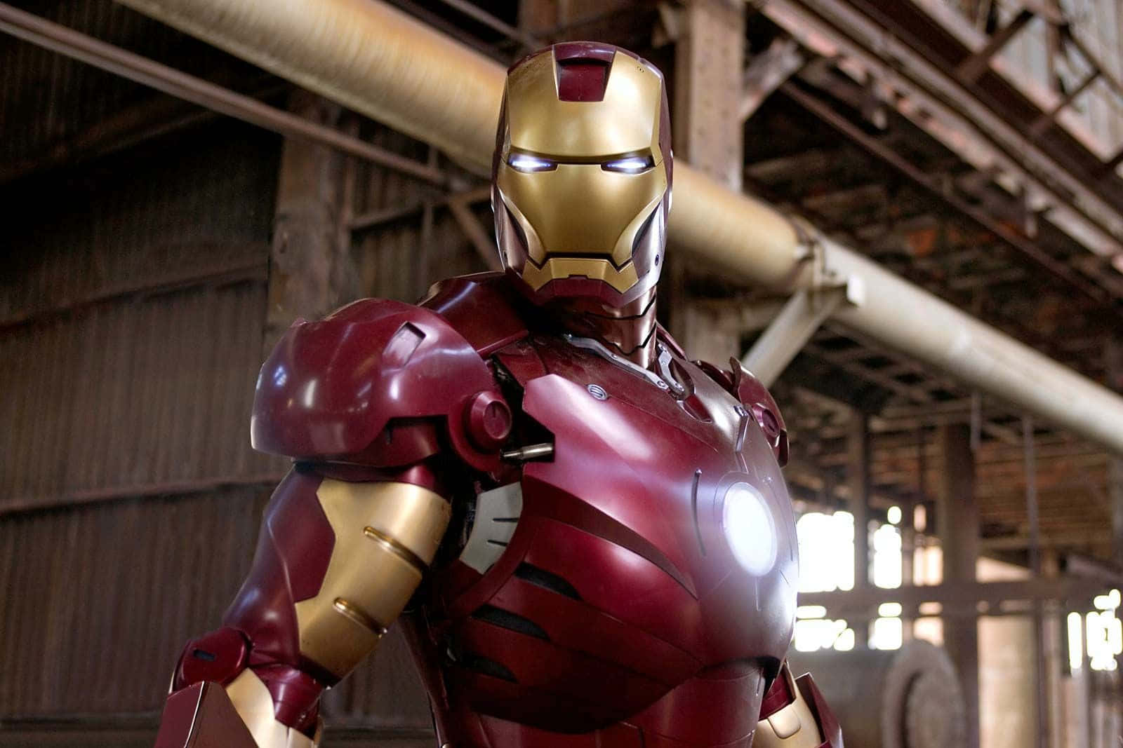 Superhero Movies 桌布 壁紙 — 鋼鐵人站在昏暗的工業倉庫中，眼睛和胸部弧形反應器發光。
