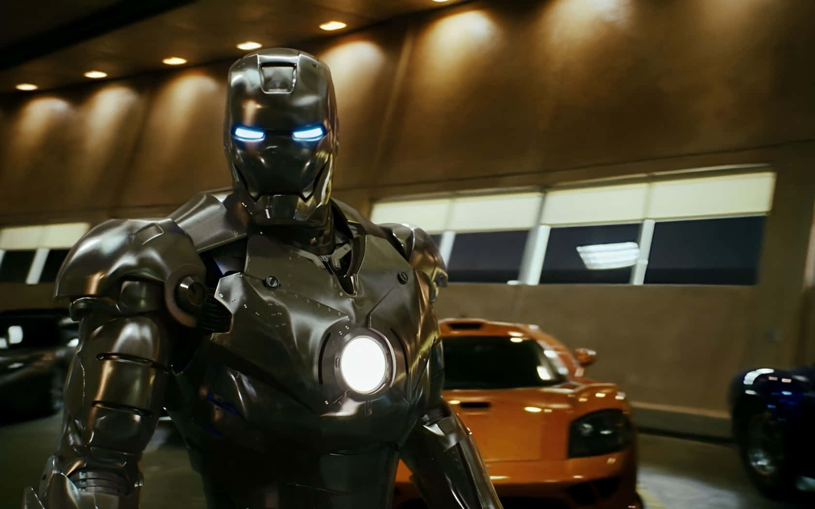 Iron Man Suit 桌布 壁紙 — 鋼鐵人站在時尚的車庫中，眼睛和胸部弧形反應爐發光，旁邊是一輛充滿活力的橙色跑車。