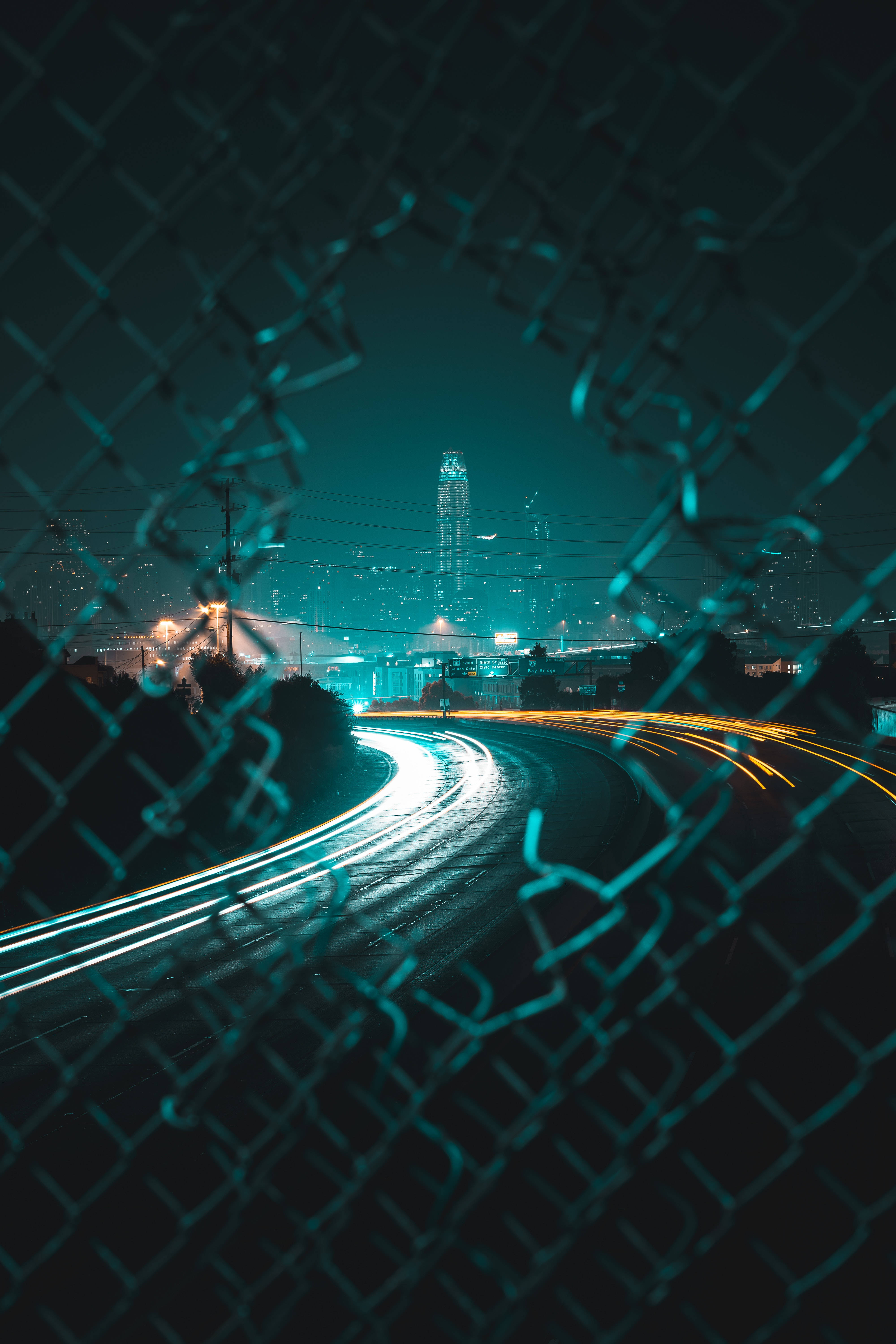 Screen 桌布 壁紙 — 鐵絲網圍欄框住了夜間蜿蜒道路上汽車發出的發光光跡，遠處是城市天際線。