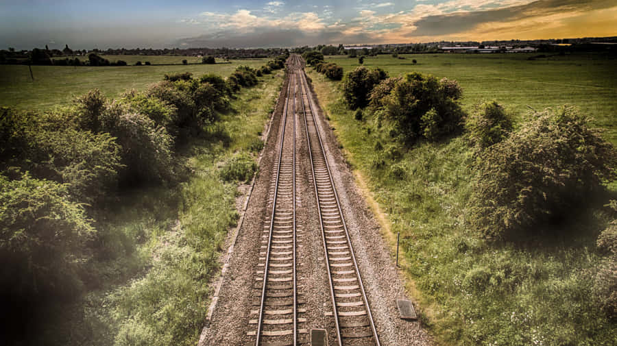 Train Tracks 桌布 壁紙 — 鐵路軌道穿過鬱鬱蔥蔥的綠色田野，在戲劇性的日落天空下延伸至遠方。