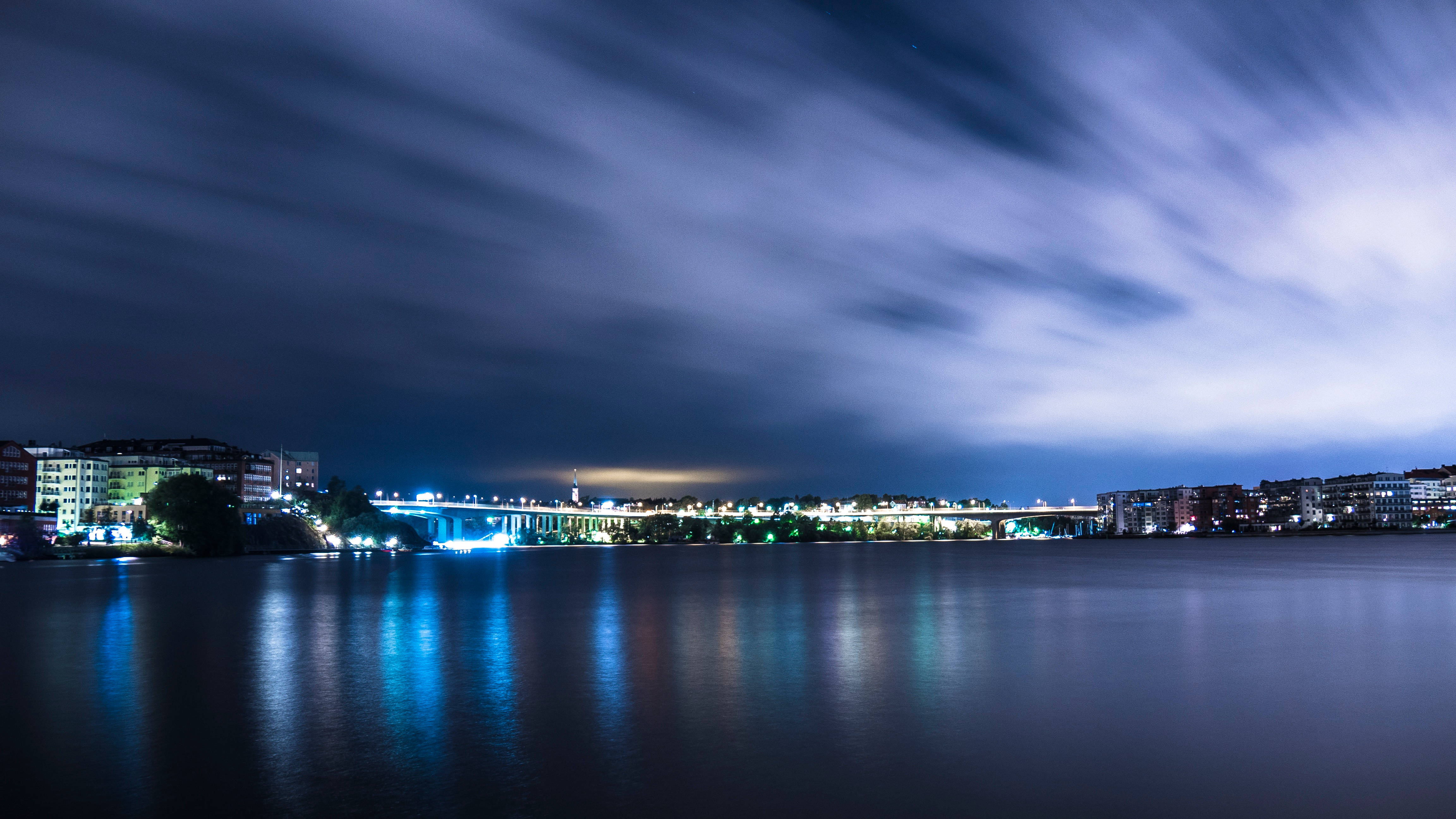 夜空 桌布 壁紙 — 長時間曝光夜景捕捉到發光的橋樑和城市燈光，在平靜的水面上反射，天空有條紋雲。