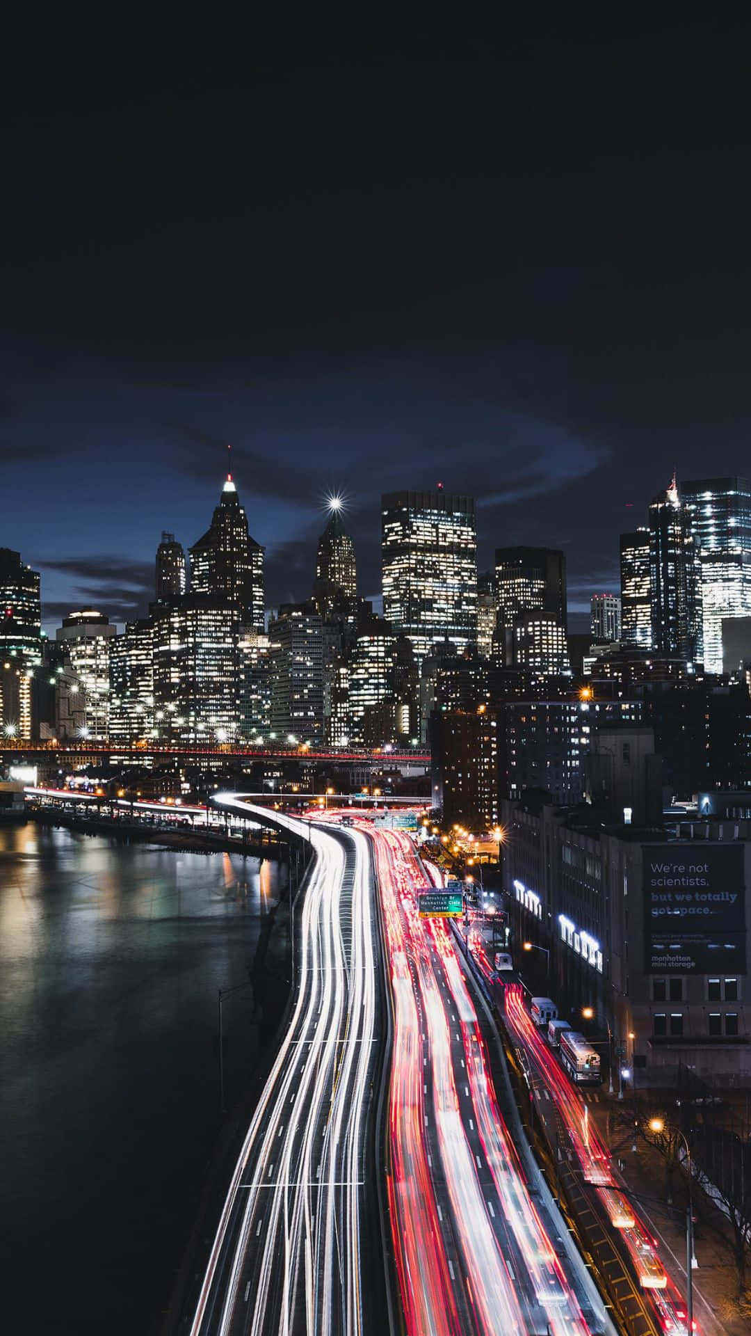 Android Ny City 桌布 壁紙 — 長時間曝光捕捉到在河濱高速公路上流動的交通所產生的發光光跡，下方是黑暗的天空，被城市天際線照亮。