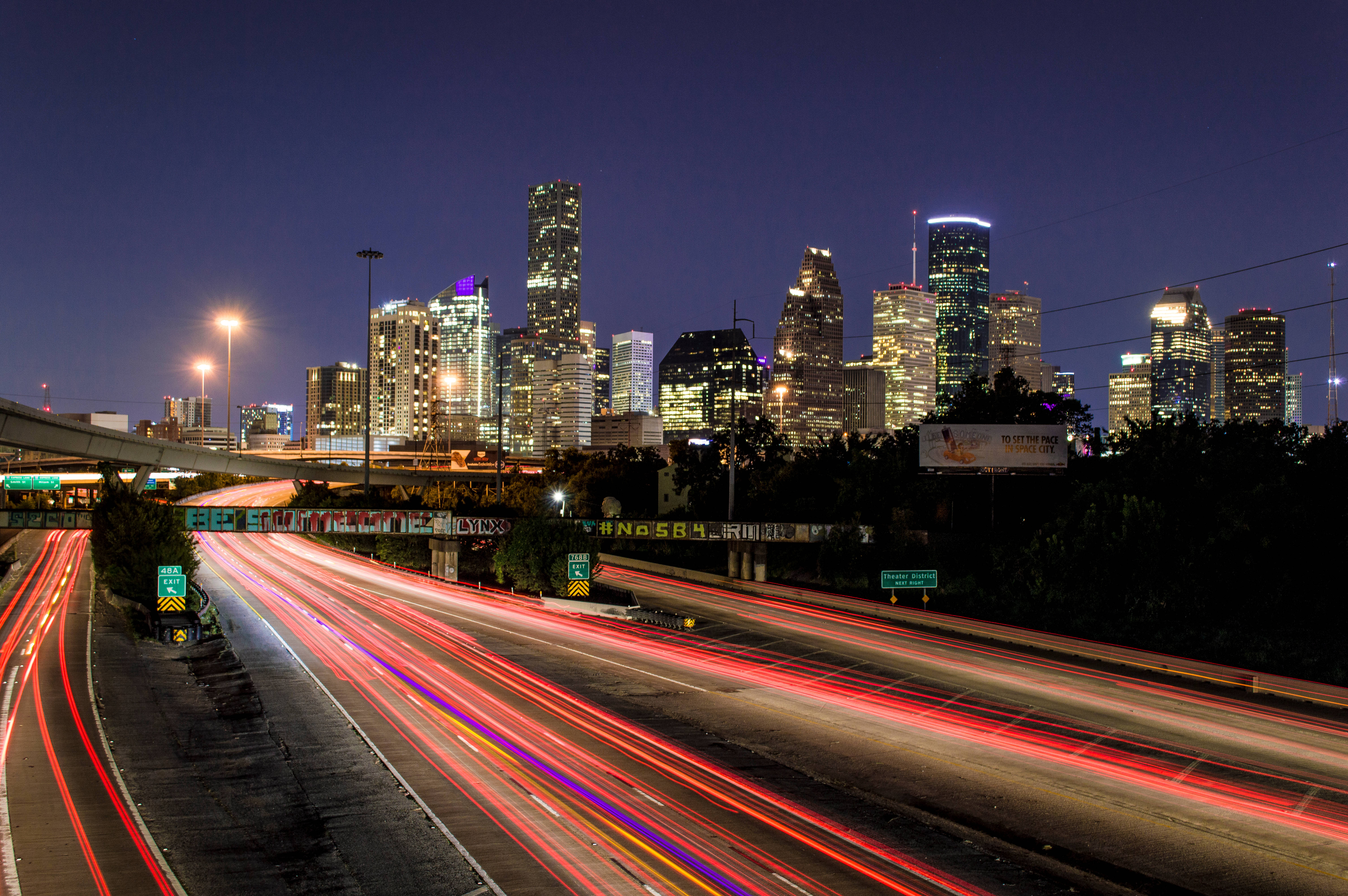 Houston 桌布 壁紙 — 長時間曝光捕捉到在發光的城市天際線下，高速公路上一道道紅色和白色車燈劃過的景象。