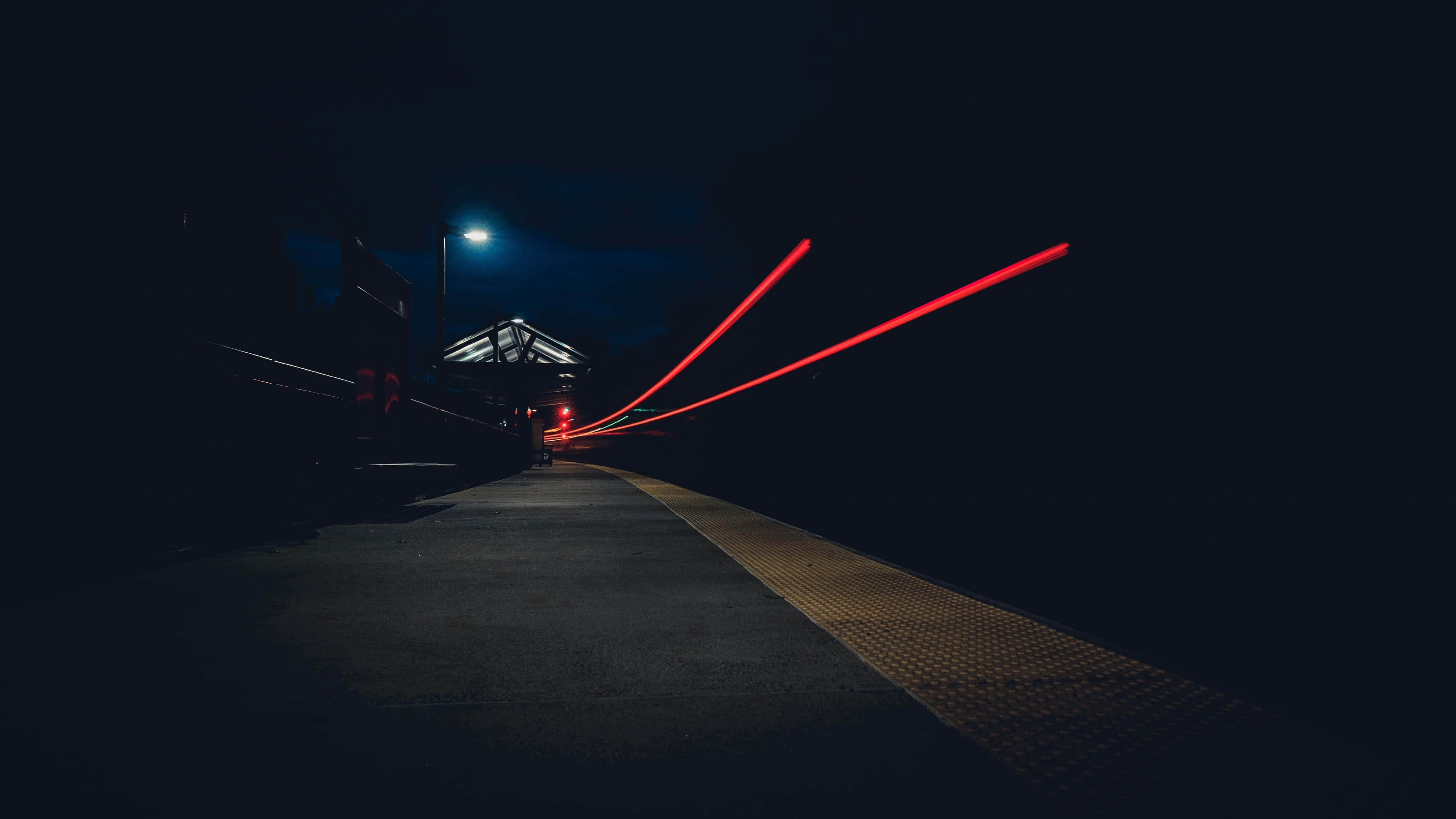 Night Street 桌布 壁紙 — 長時間曝光捕捉到在黑暗的火車站月台上，在一盞發光路燈下，鮮紅色光軌劃過的景象。