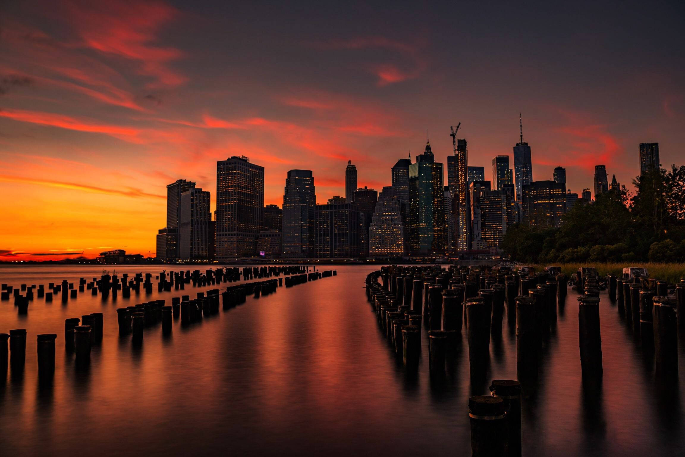 Nyc Aesthetic 桌布 壁紙 — 長時間曝光捕捉到城市天際線上熾熱的日落，發光的摩天大樓倒映在平靜的水面上，並以風化的木樁為框。