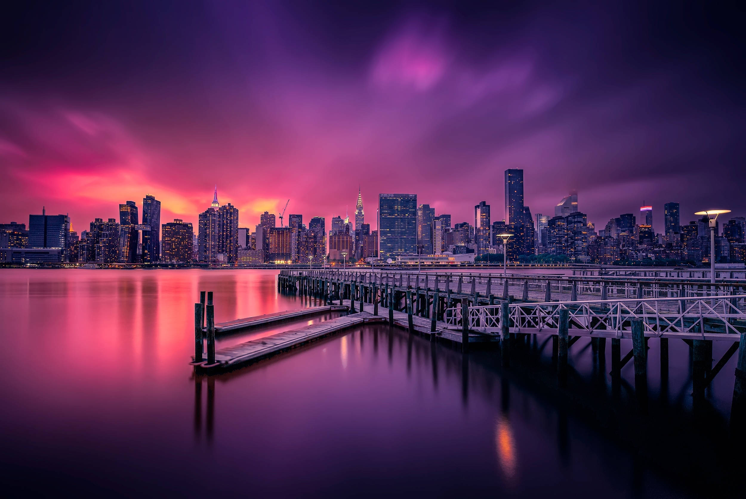 New York Skyline 桌布 壁紙 — 長時間曝光捕捉到城市天際線上空充滿活力的紫色和粉紅色日落，倒映在平靜的水面上，前景是木製碼頭。