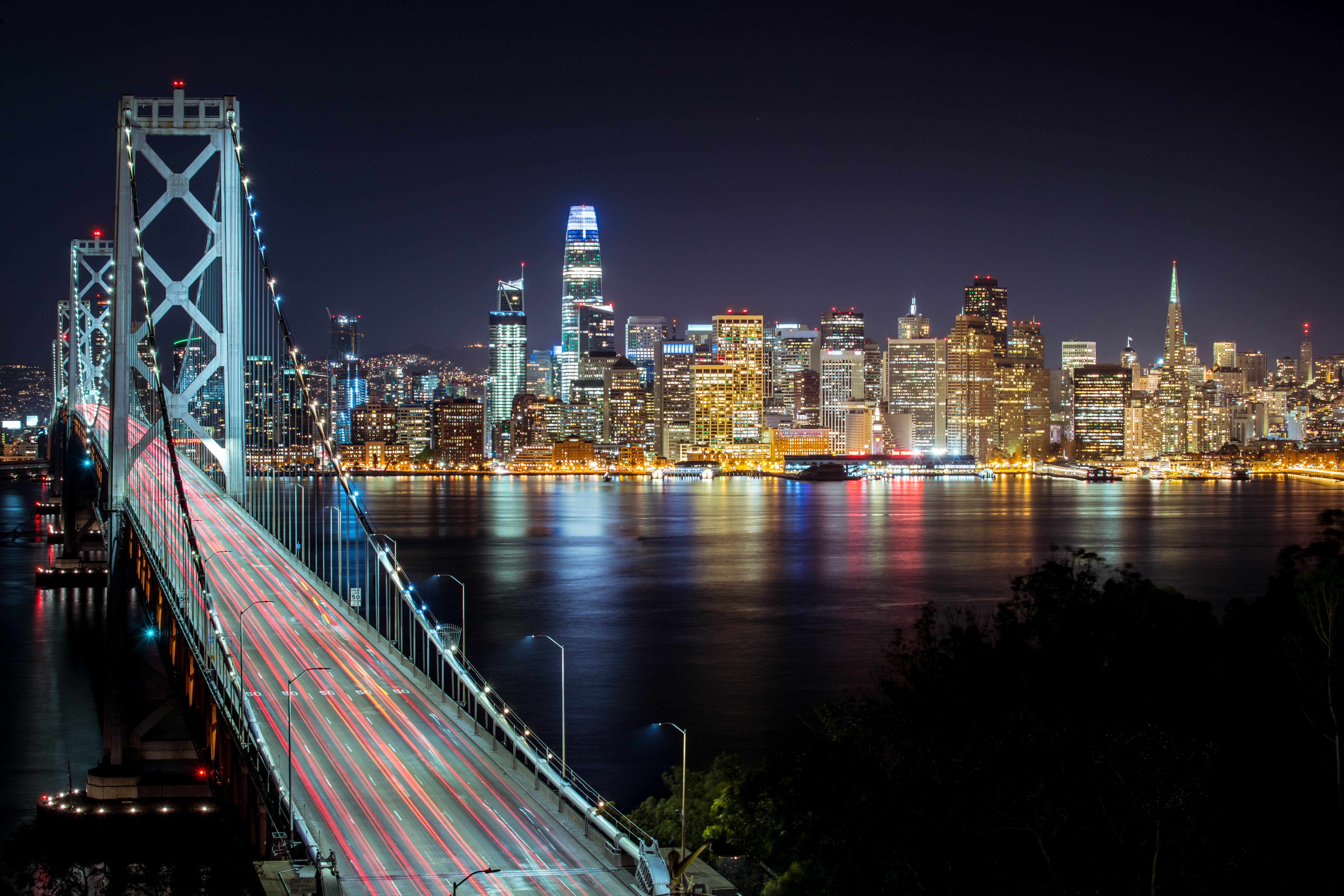 San Francisco Phone 桌布 壁紙 — 長時間曝光捕捉到夜間橋上發光的燈光軌跡，以及水中倒映的充滿活力的城市天際線。