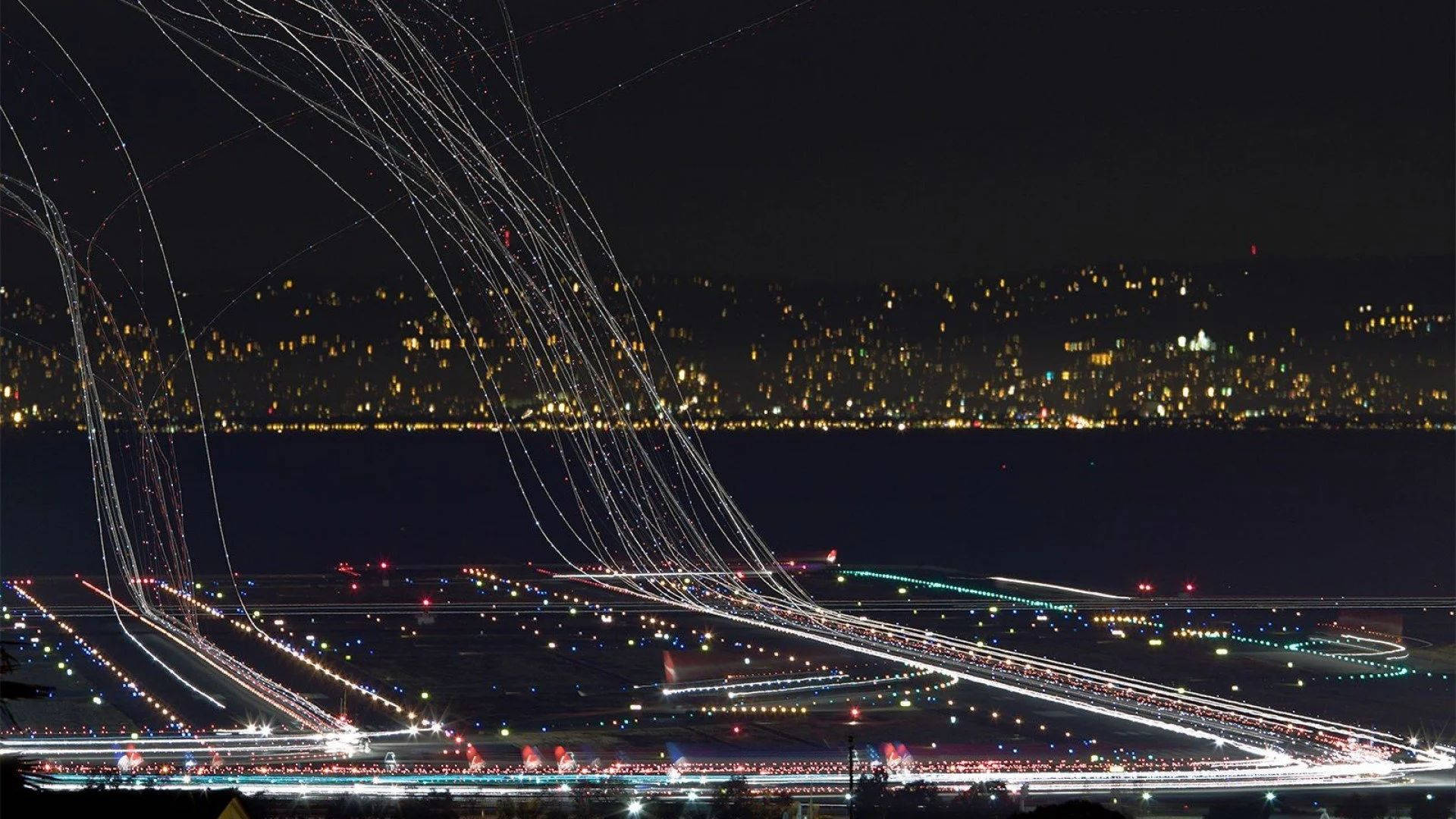 Runway 桌布 壁紙 — 長時間曝光捕捉到夜間機場上空飛機燈光的軌跡，遠方是發光的城市景觀。