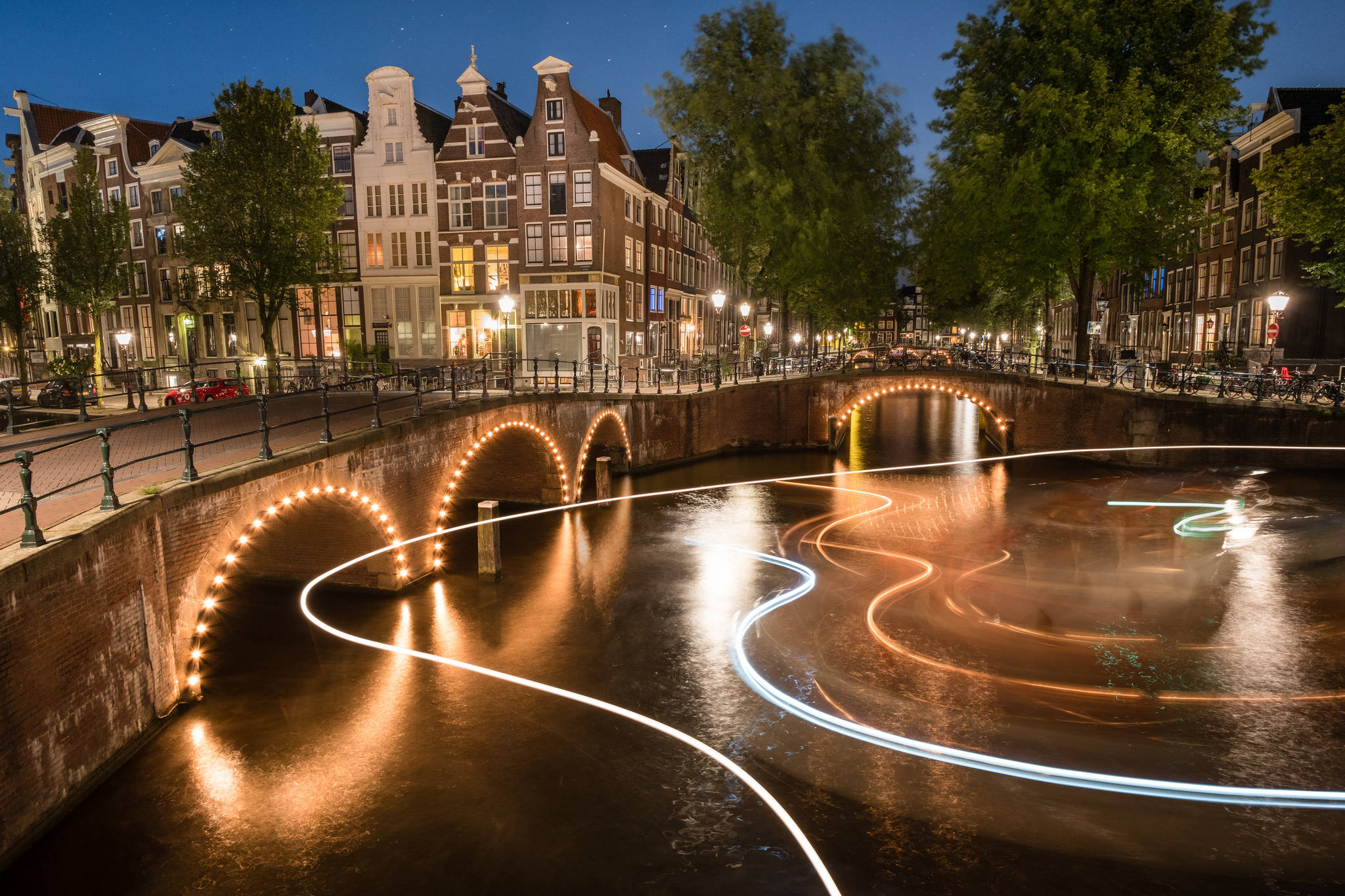 Netherlands 桌布 壁紙 — 長時間曝光捕捉到夜間歐洲城市歷史拱橋下運河上船隻舞動的發光軌跡。