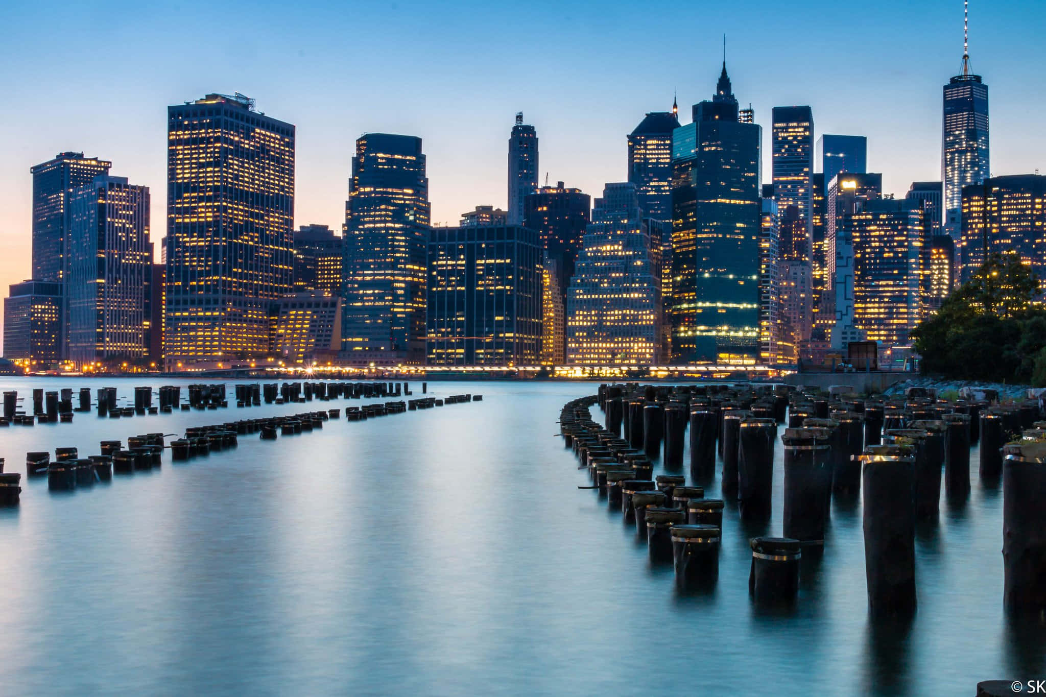 New York City Landscape 桌布 壁紙 — 長時間曝光捕捉到暮色中平靜水面上閃爍的摩天大樓倒影，前景是一排排風化的木樁。