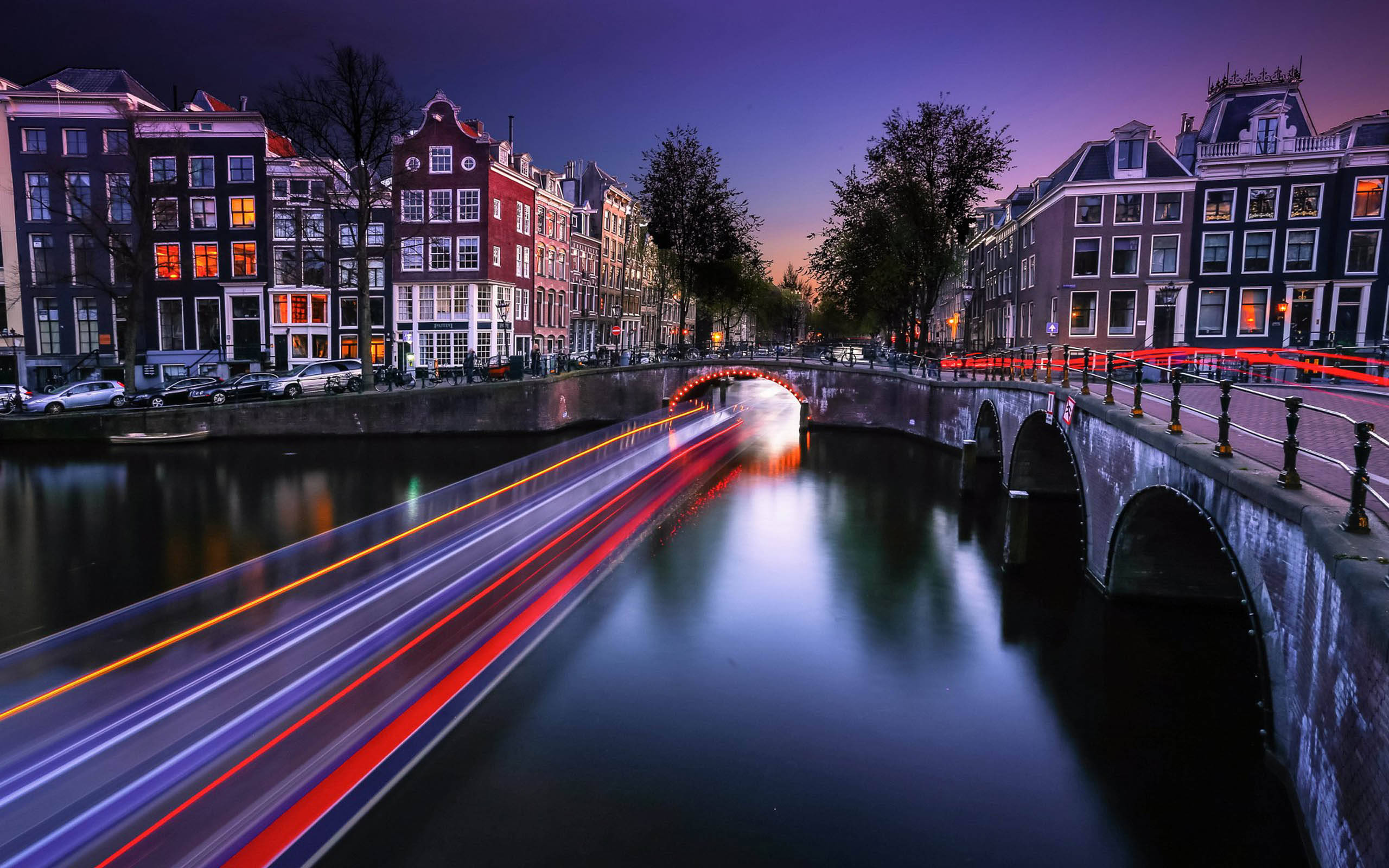 Netherlands 桌布 壁紙 — 長時間曝光捕捉到阿姆斯特丹運河上船隻的光跡，暮色中歷史建築溫暖發光。