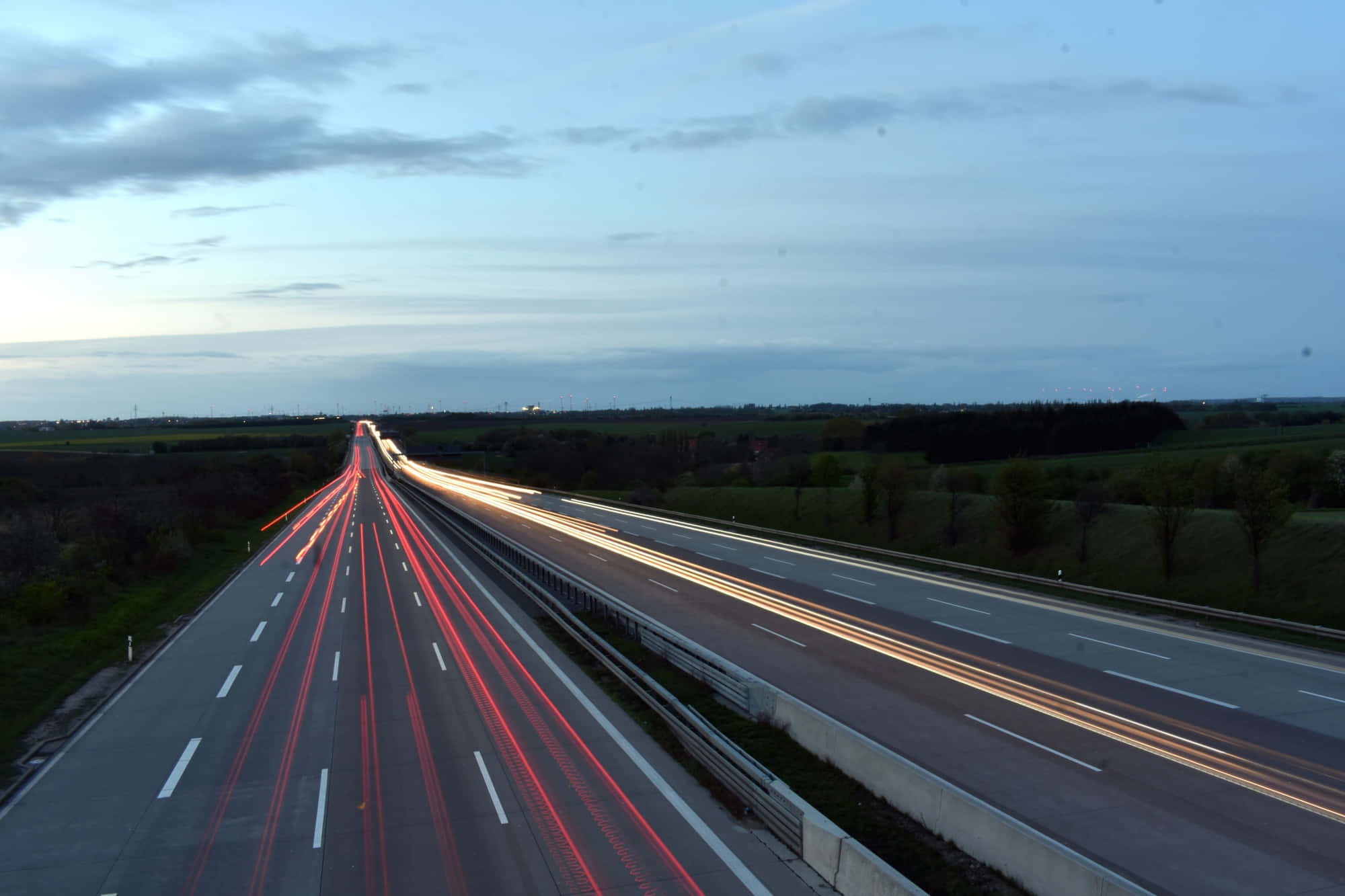 Highways 桌布 壁紙 — 長時間曝光捕捉到黃昏時分高速公路上車流的紅色和白色光線，天空多雲，田野翠綠。