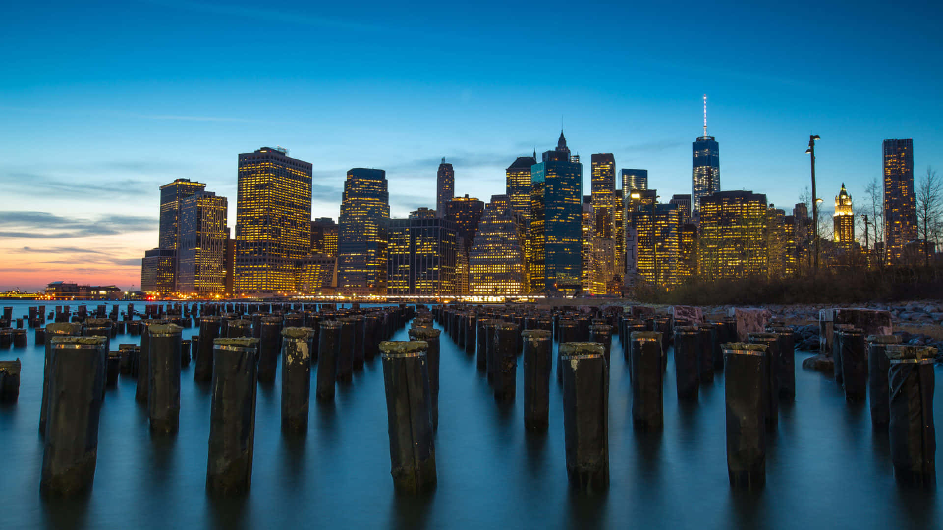 New York City 4K Ultra HD 桌布 壁紙 — 長時間曝光捕捉到黃昏時發光的摩天大樓倒映在平靜的水面上，前景有風化的樁柱。