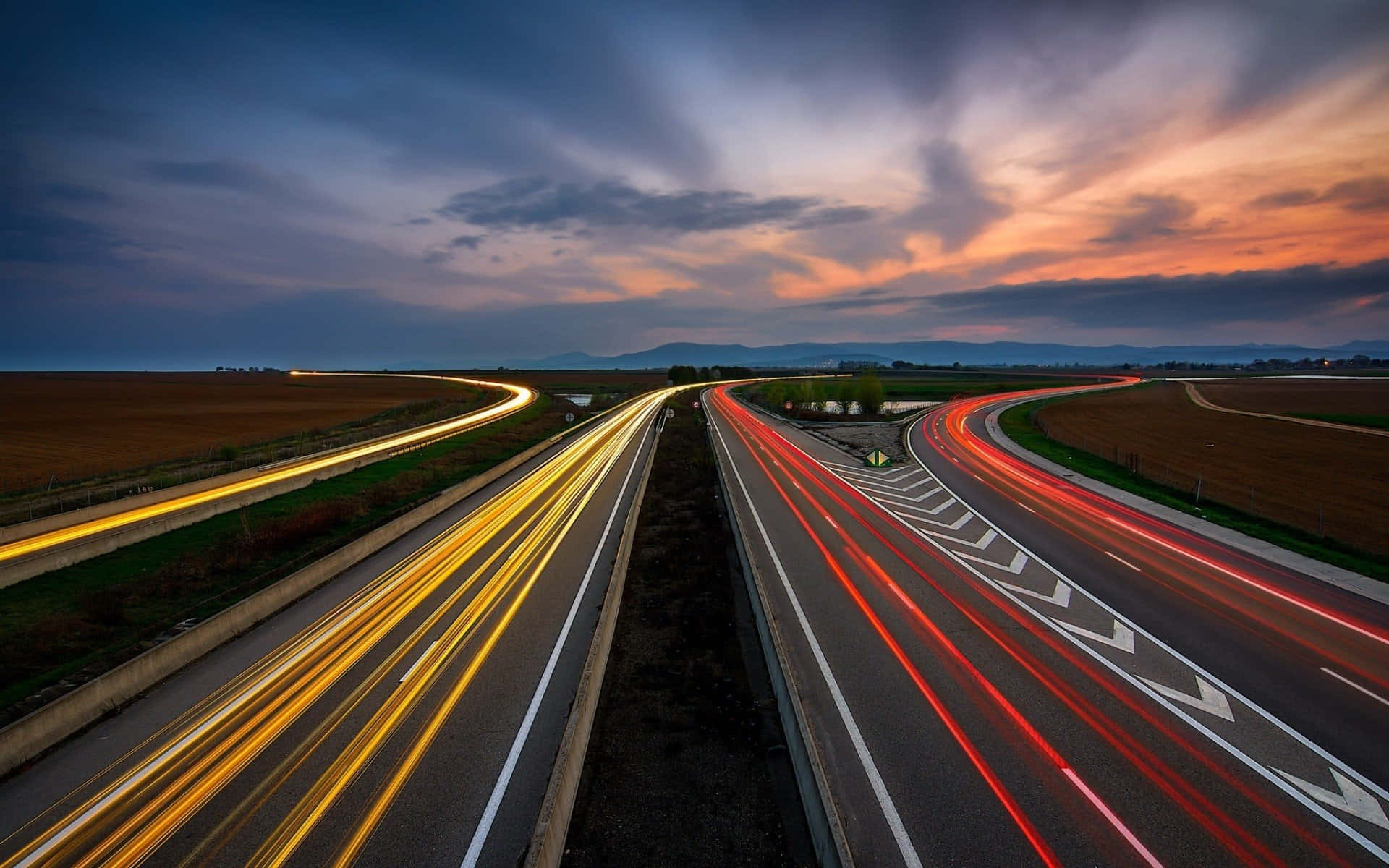 Highways 桌布 壁紙 — 長時間曝光捕捉到黃昏時高速公路上汽車燈光的流線，伴隨戲劇性的雲層和遠山。