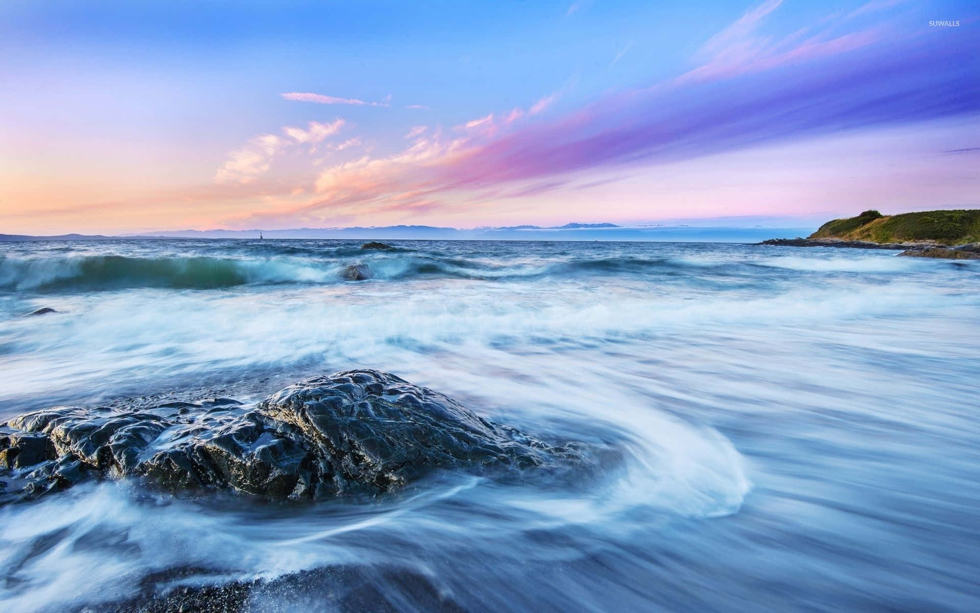 Ocean Waves 桌布 壁紙 — 長時間曝光的海景捕捉到平滑的藍色波浪在深色岩石周圍旋轉，下方是充滿活力的紫色和橙色日落天空。