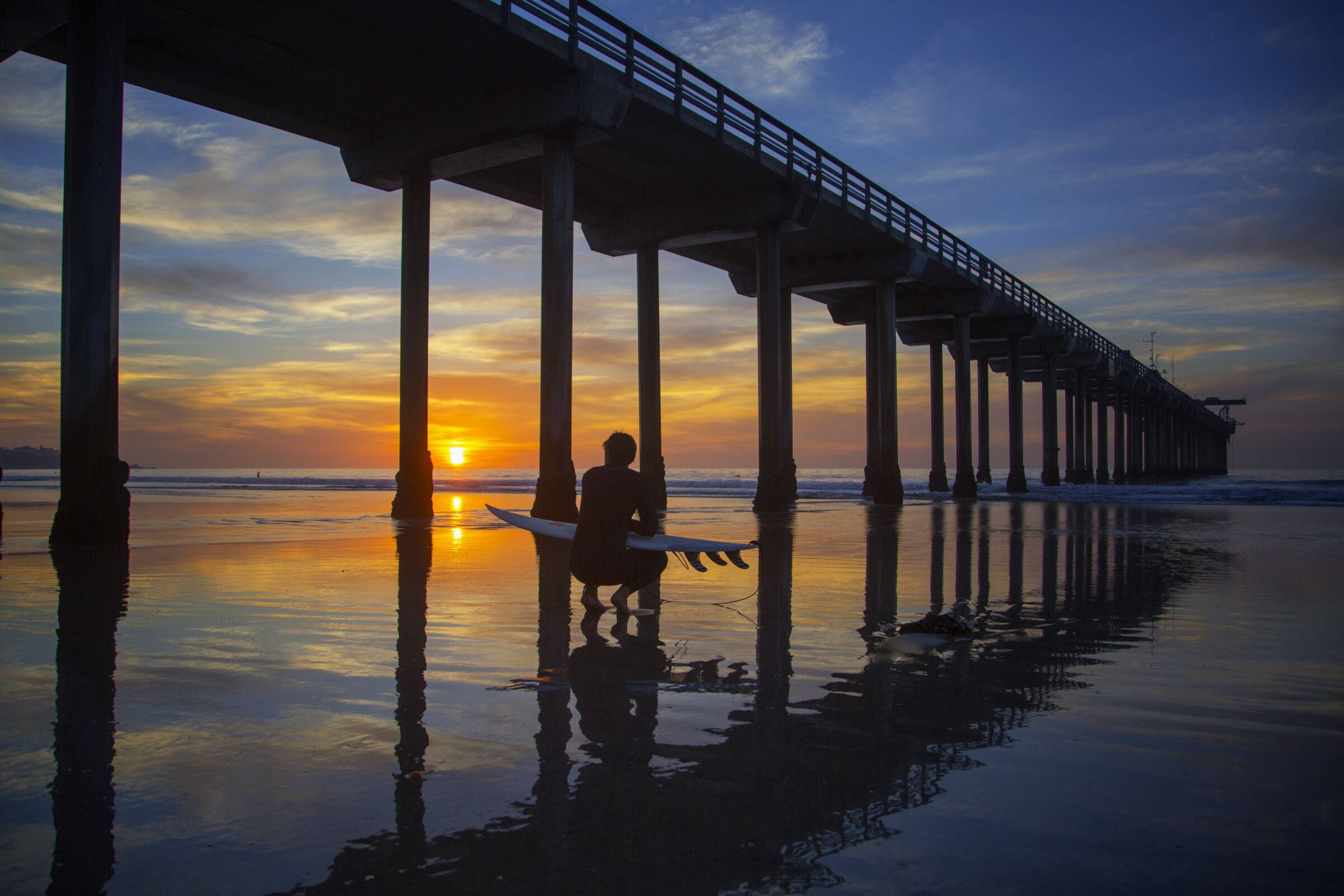美麗 桌布 壁紙 — 長碼頭下，一名衝浪者蹲伏，手持衝浪板，金色夕陽映照在濕沙上。