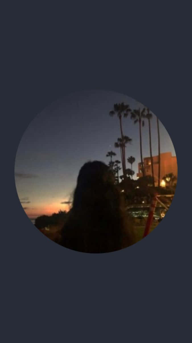 Instagram Profile 桌布 壁紙 — 長髮人物的剪影，凝視著由棕櫚樹和發光建築物框住的黃昏天空。