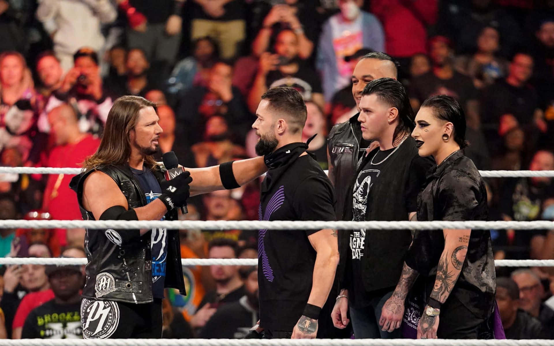 WWE 桌布 壁紙 — 長髮摔角手拿著麥克風，在擂台燈光下與對手對峙，觀眾熱切觀看。