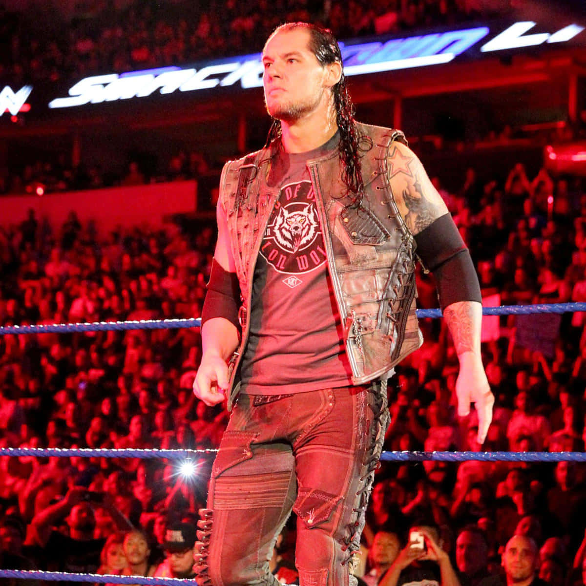 WWE 桌布 壁紙 — 長黑髮及紋身的摔角手站在紅色燈光下的摔角擂台上，身後有歡呼的人群。