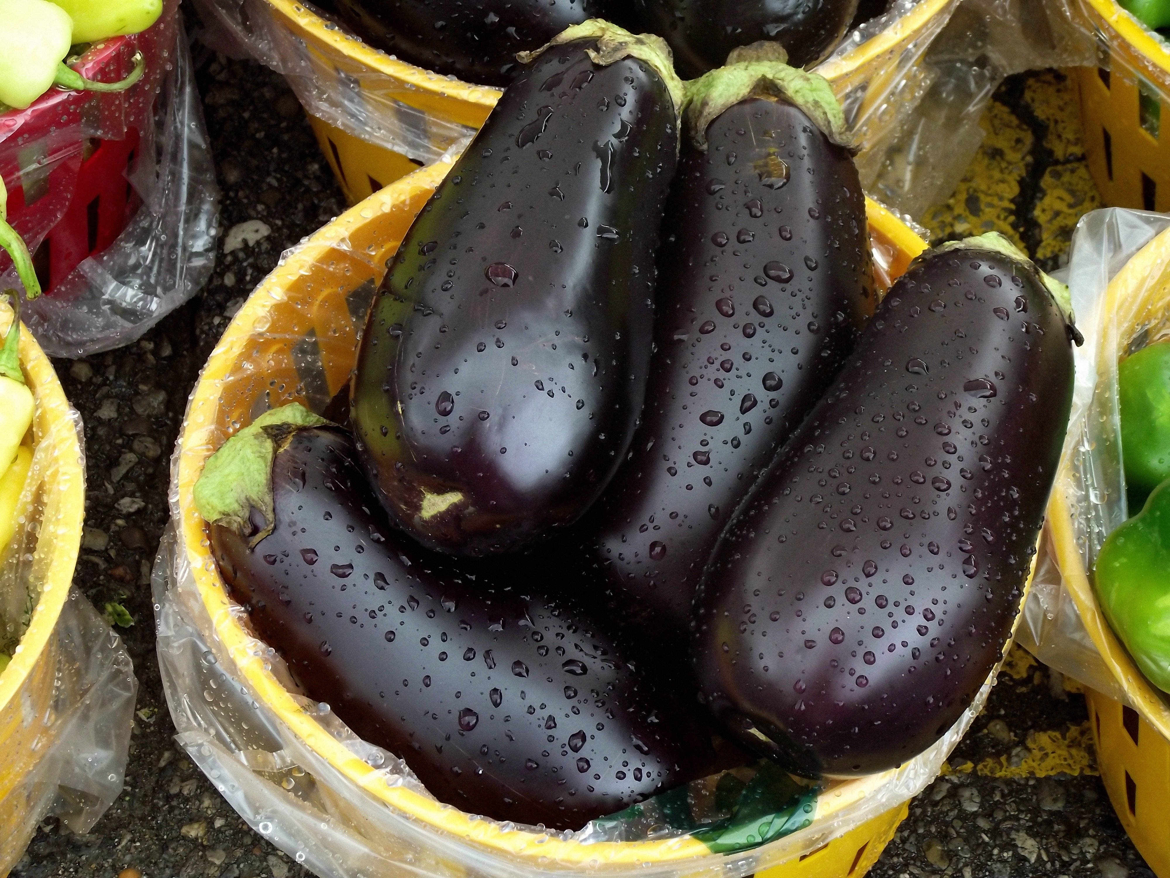 Fruits 桌布 壁紙 — 閃亮深紫色茄子帶有水珠，放在一個黃色籃子裡，在充滿活力的戶外市場攤位上。