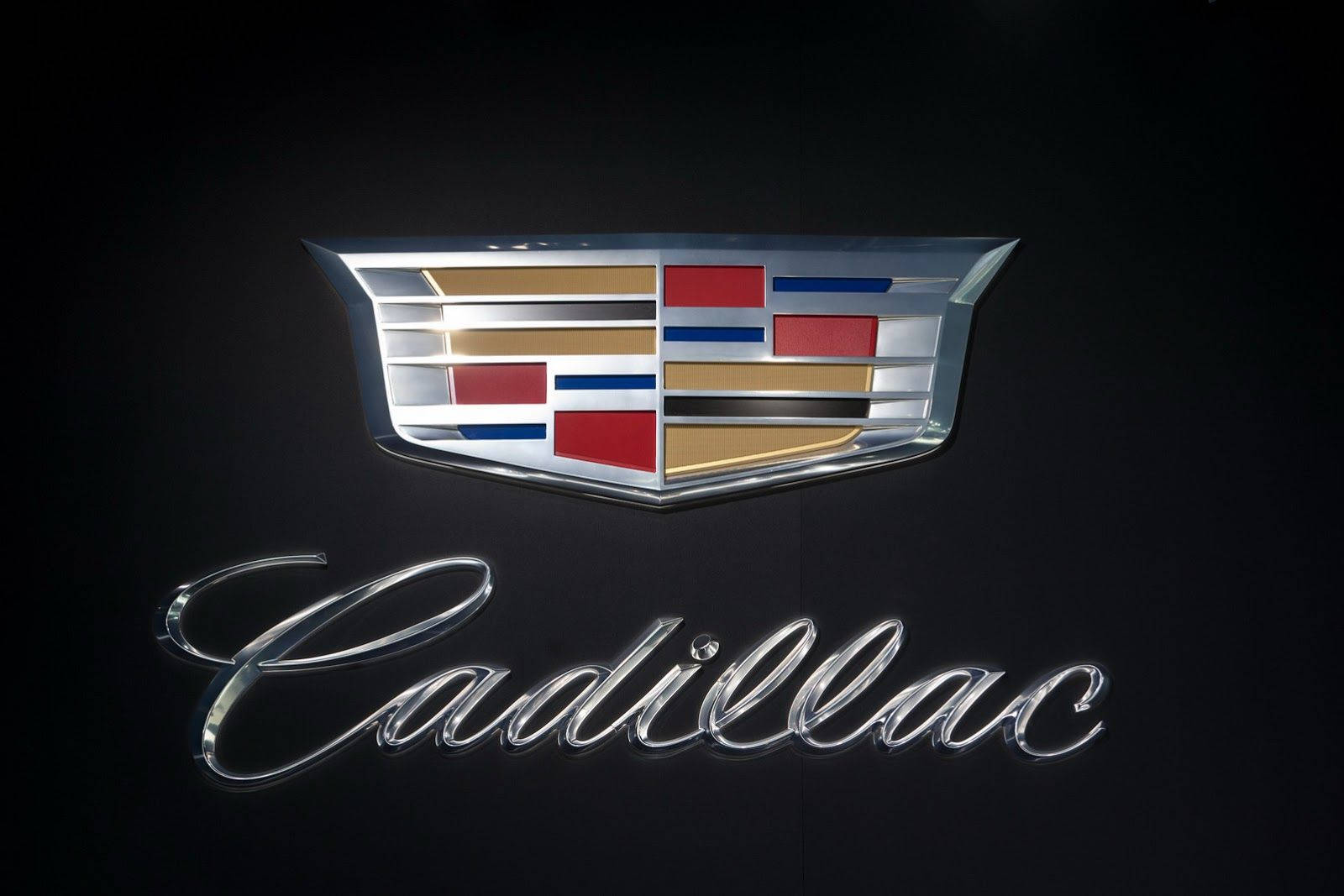 Cadillac 桌布 壁紙 — 閃亮的凱迪拉克徽章，帶有紅色、金色和藍色條紋，在黑色背景上閃耀，下方有草書標誌。