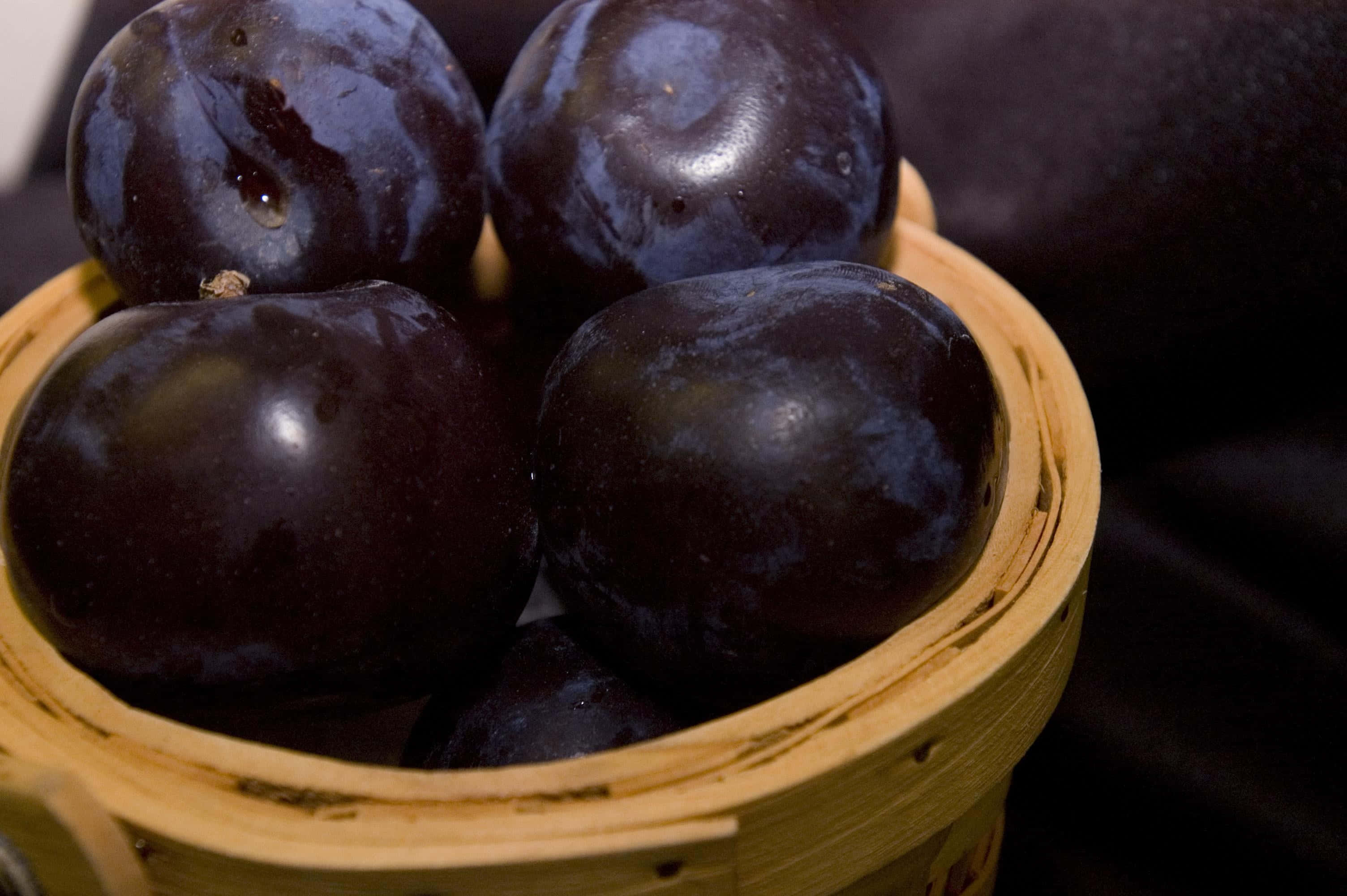 Purple Plums 桌布 壁紙 — 閃亮的深紫色李子放在編織竹籃中，背景柔和模糊。