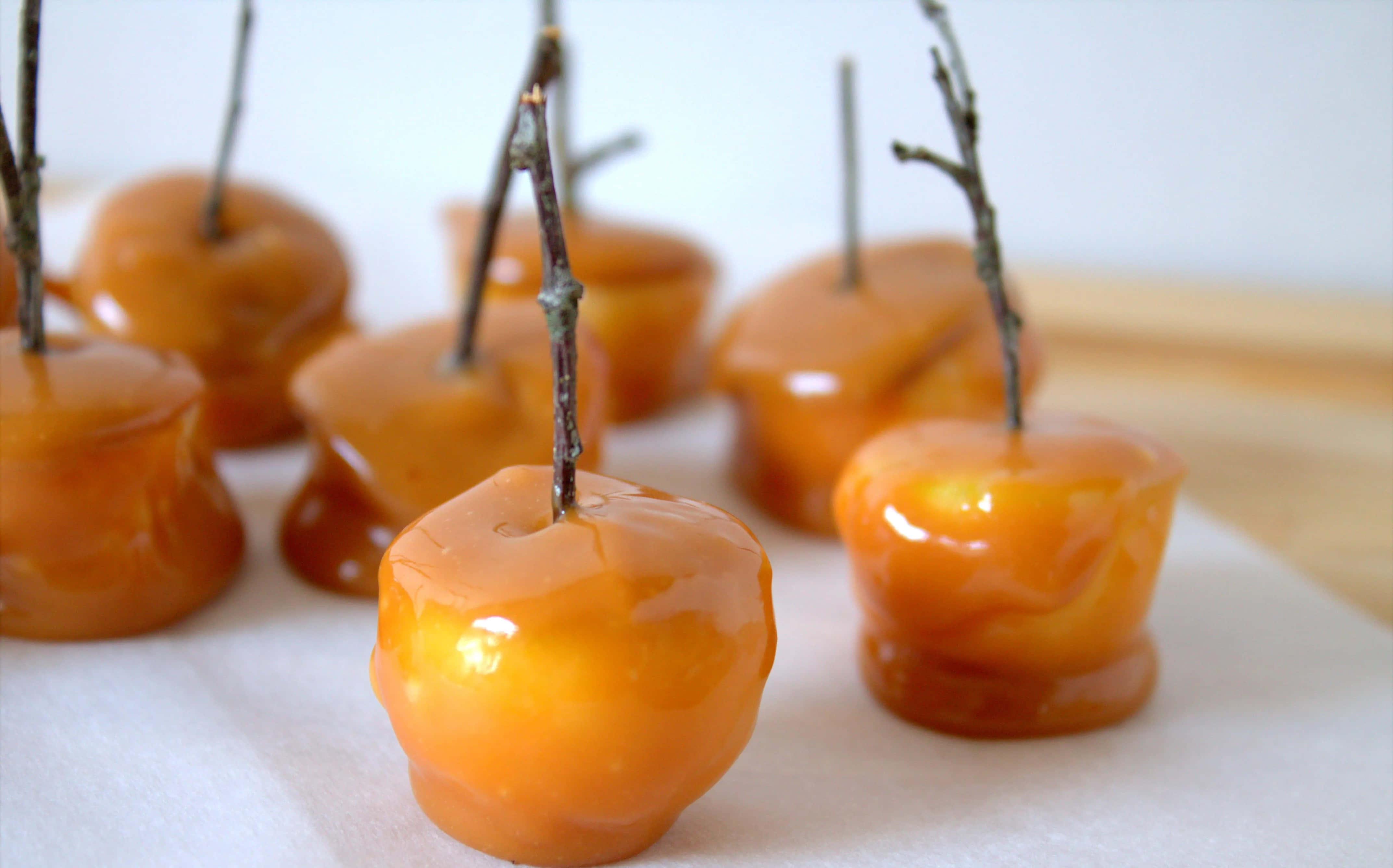 Caramel Apples 桌布 壁紙 — 閃亮的焦糖蘋果帶有樹枝莖，放在烘焙紙上，襯以柔和的白色背景。