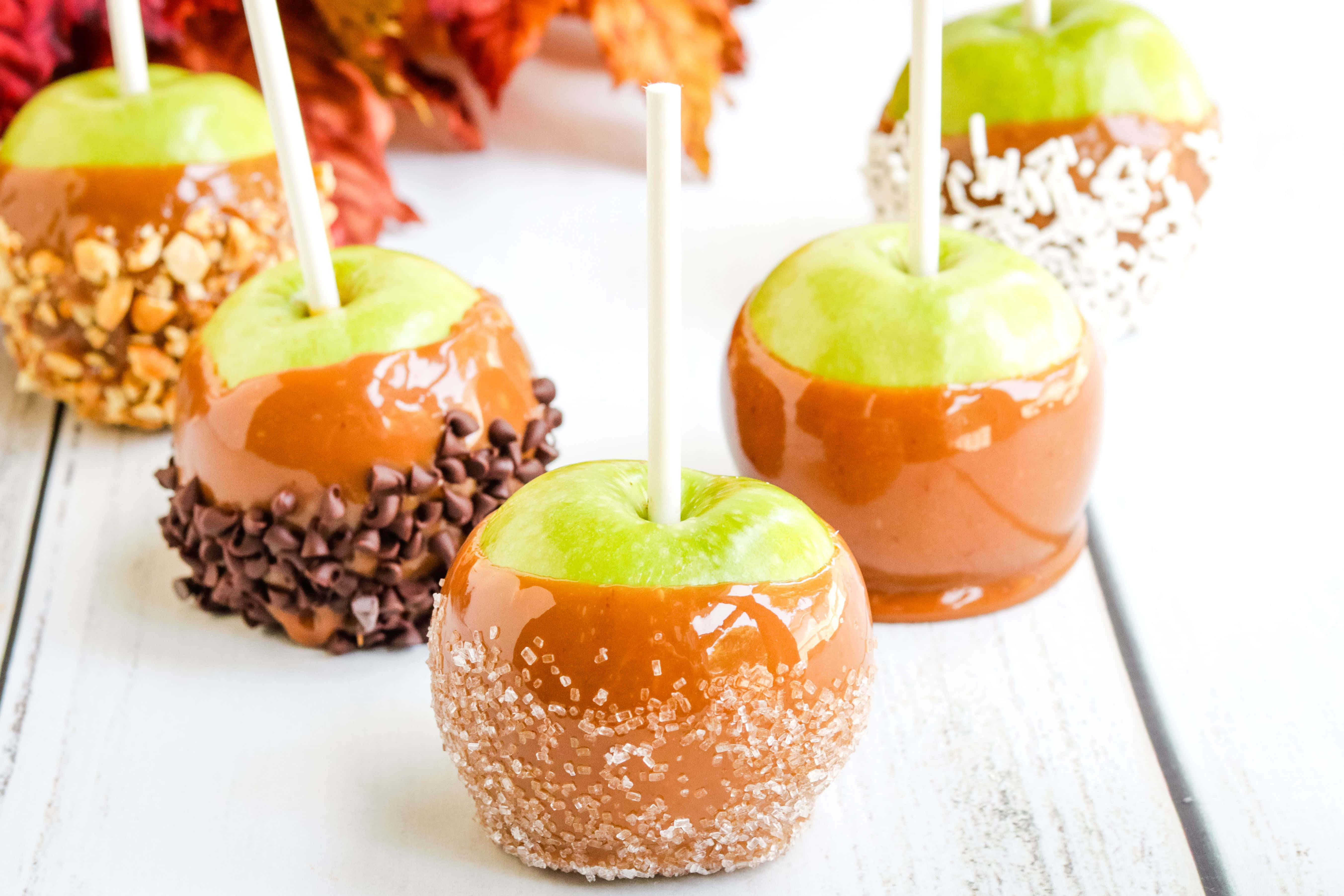 Caramel Apples 桌布 壁紙 — 閃亮的焦糖蘋果裹上堅果、巧克力、鹽和椰子，放在白色木質表面上，背景有秋葉。
