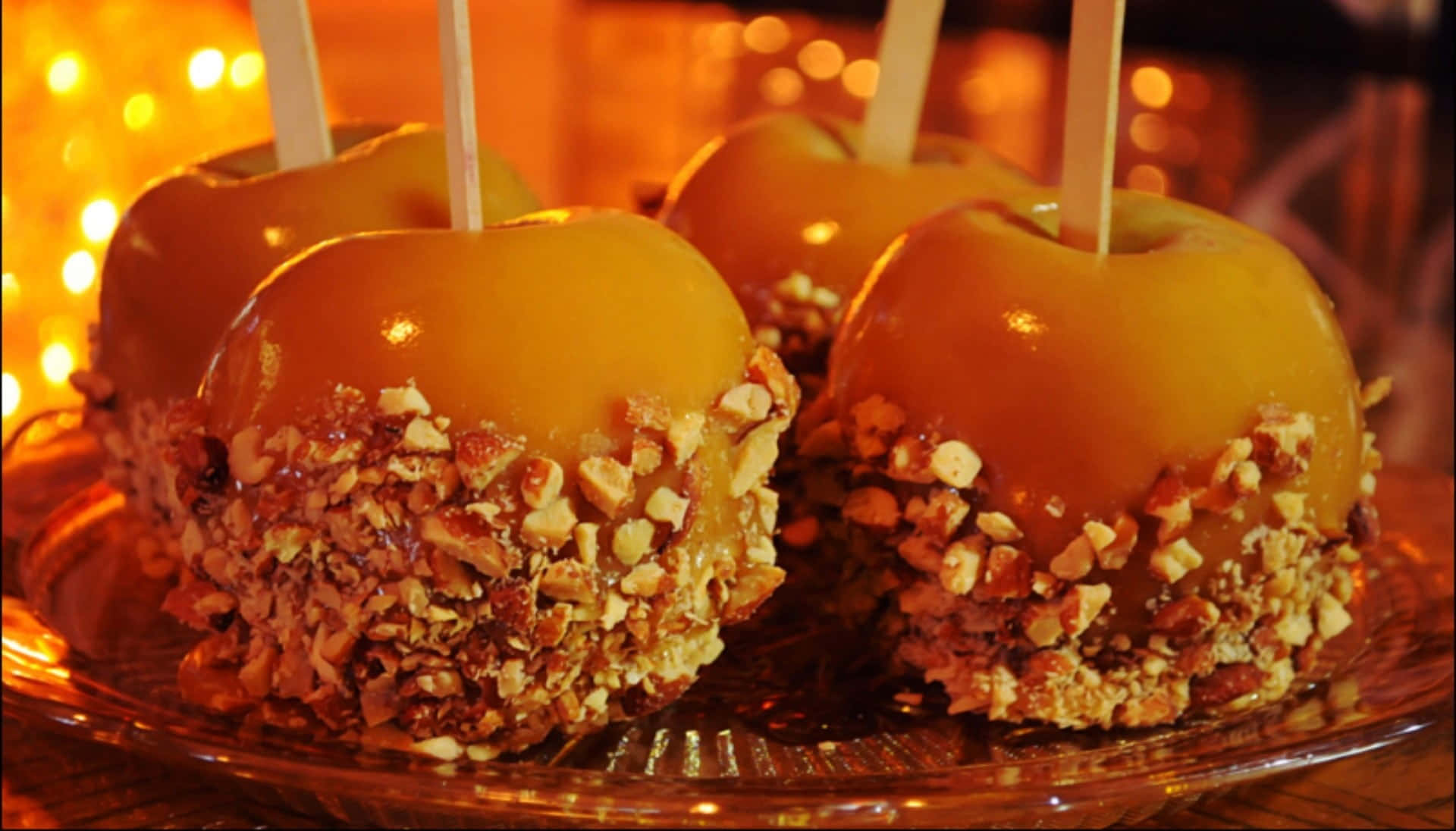 Caramel Apples 桌布 壁紙 — 閃亮的焦糖蘋果裹上碎堅果，放在玻璃盤上，在溫暖的串燈下。