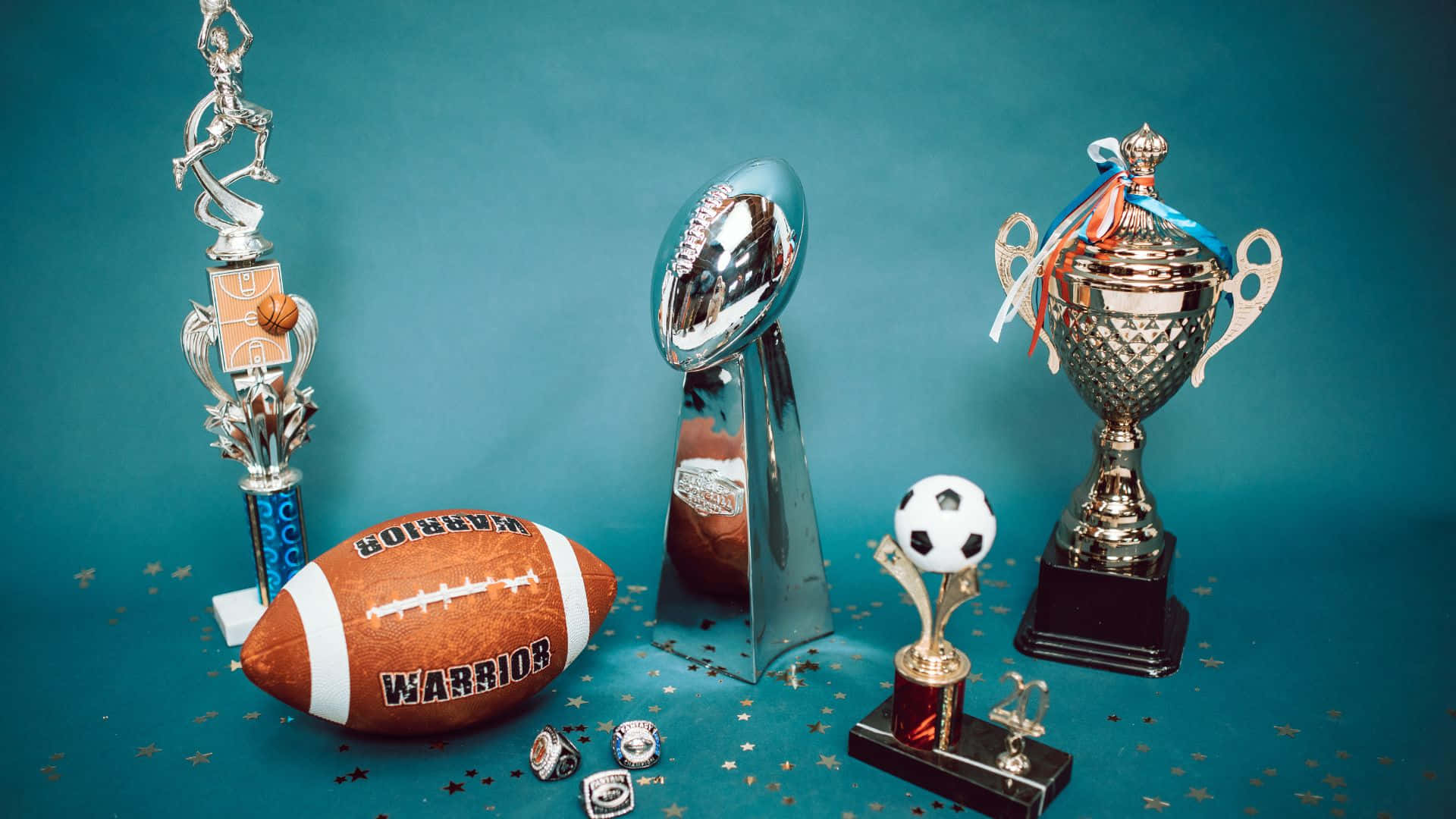 Best Sports 桌布 壁紙 — 閃亮的獎盃和運動球類，如足球和英式足球，排列在帶有亮粉的藍綠色背景上。
