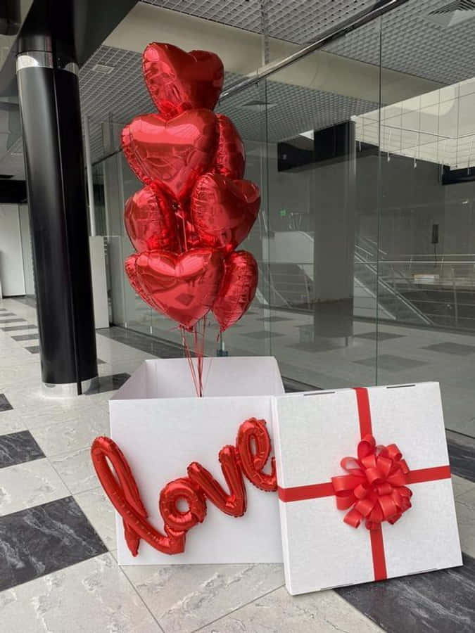 情人節 桌布 壁紙 — 閃亮的紅色心形氣球和「love」字母氣球從玻璃牆附近的格子地板上的白色禮品盒中升起。