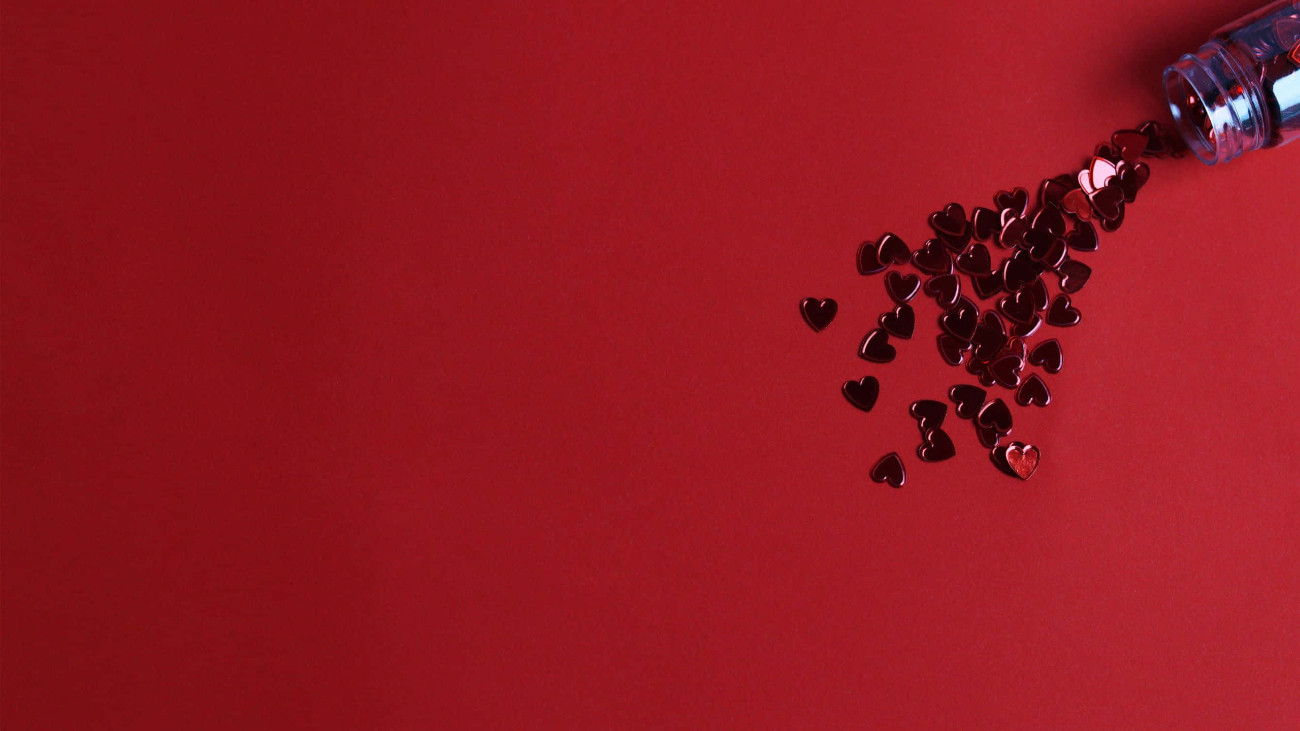 Red Color 桌布 壁紙 — 閃亮的紅色心形紙屑從透明瓶子中灑落到純紅色表面上，形成極簡的平鋪構圖。