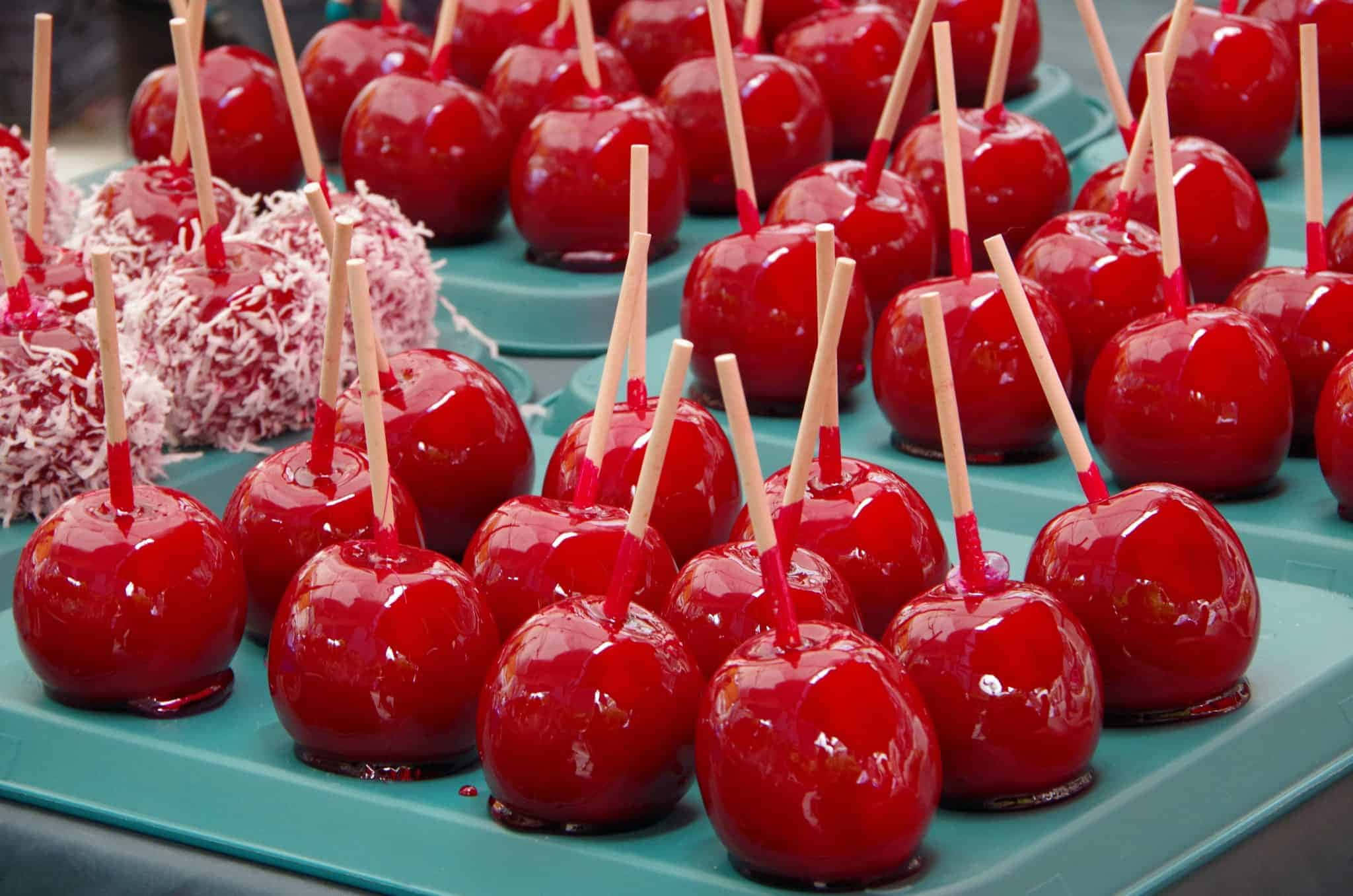 Candy Apples 桌布 壁紙 — 閃亮的紅色焦糖蘋果串在棒上，整齊排列在青綠色托盤上，部分裹上白色椰子片。