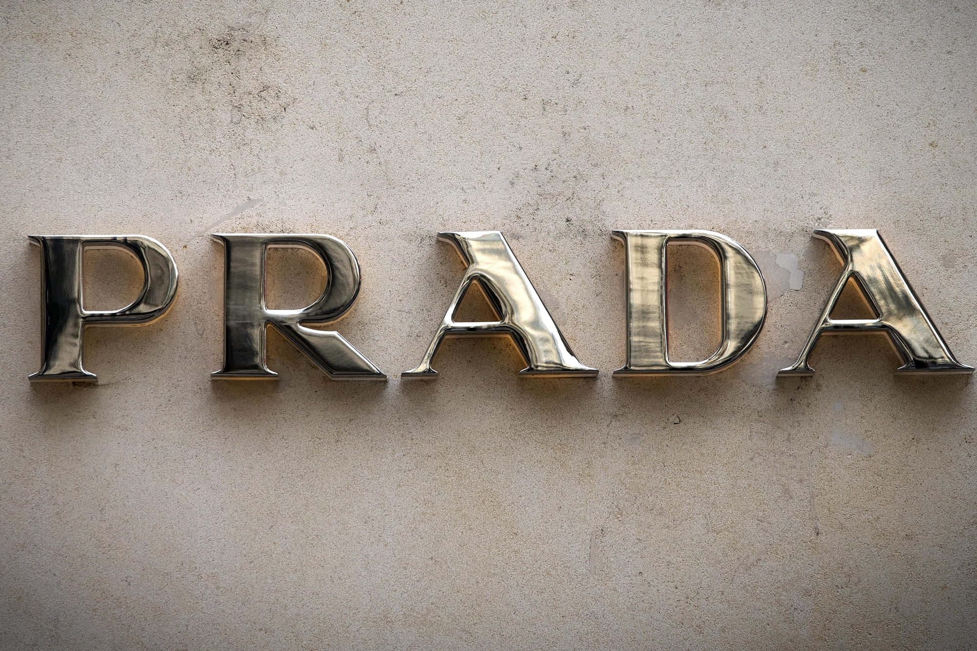 Prada 桌布 壁紙 — 閃亮的金屬Prada字母安裝在紋理米色牆上，帶有柔和環境照明。