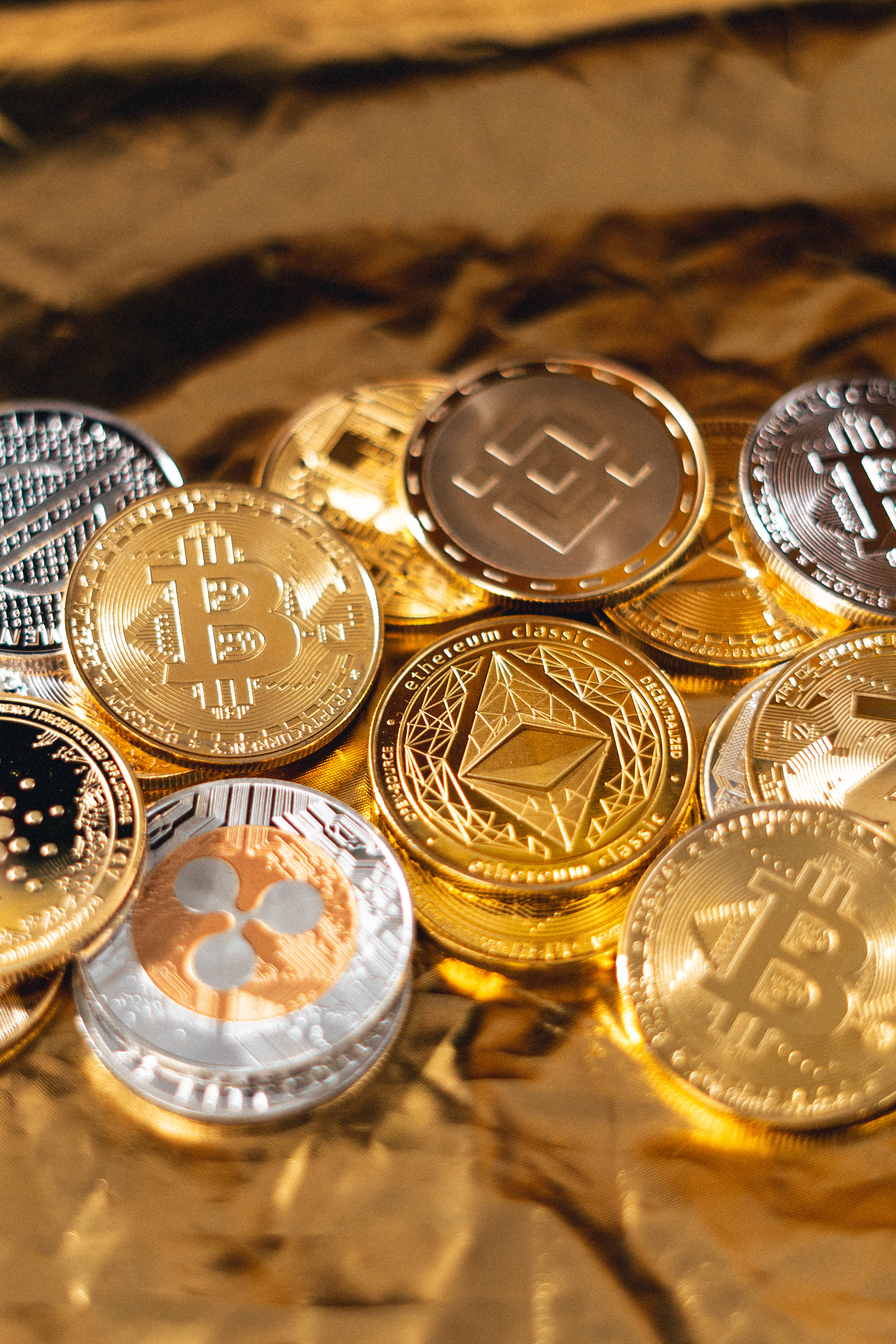 Cryptocurrency 桌布 壁紙 — 閃亮的金色和銀色加密貨幣硬幣，如比特幣和以太幣，在溫暖的光線下放在皺巴巴的金箔上。