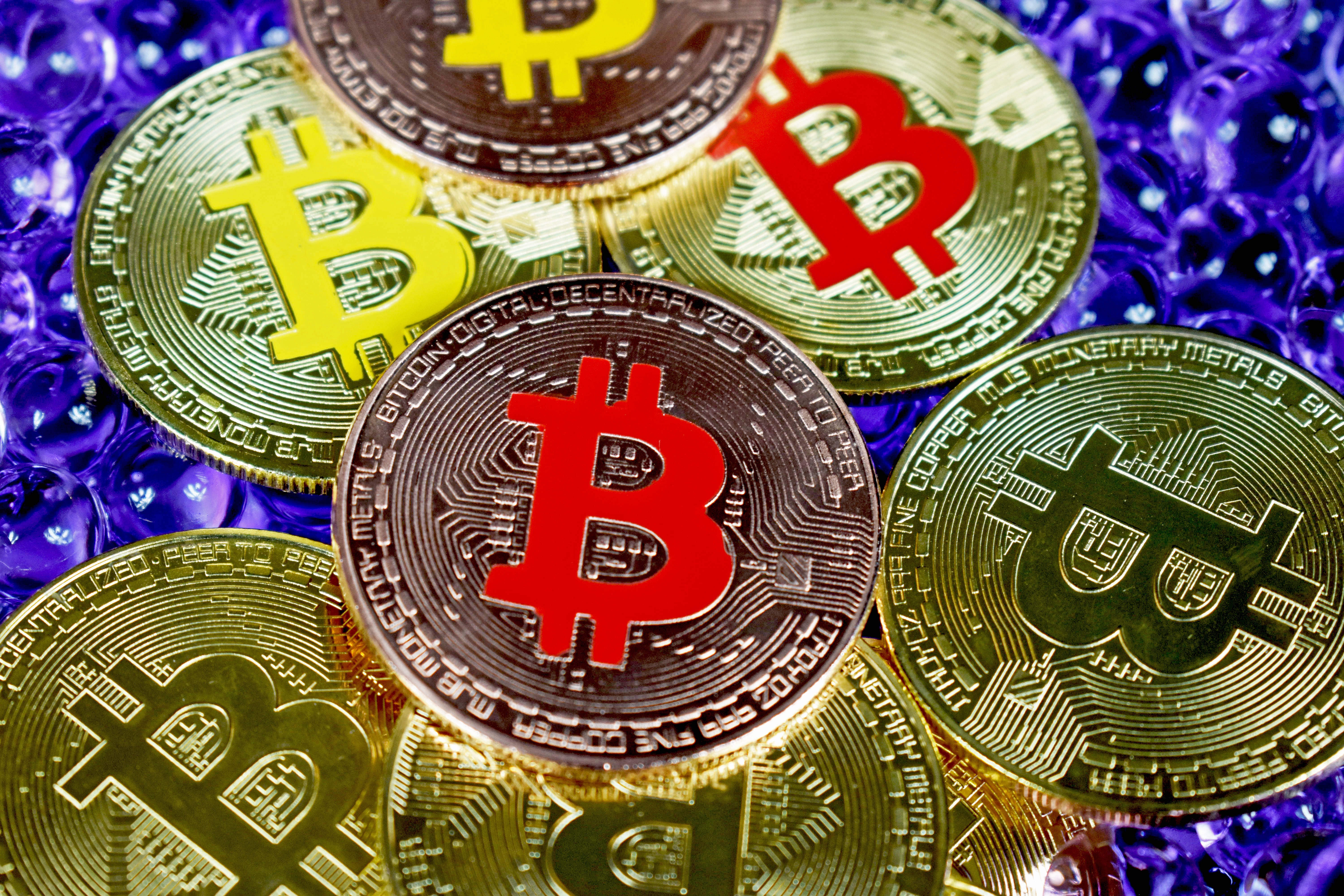 比特幣 桌布 壁紙 — 閃亮的金色和銀色比特幣硬幣，帶有紅色和黃色的「B」標誌，放在紫色泡泡紙上。