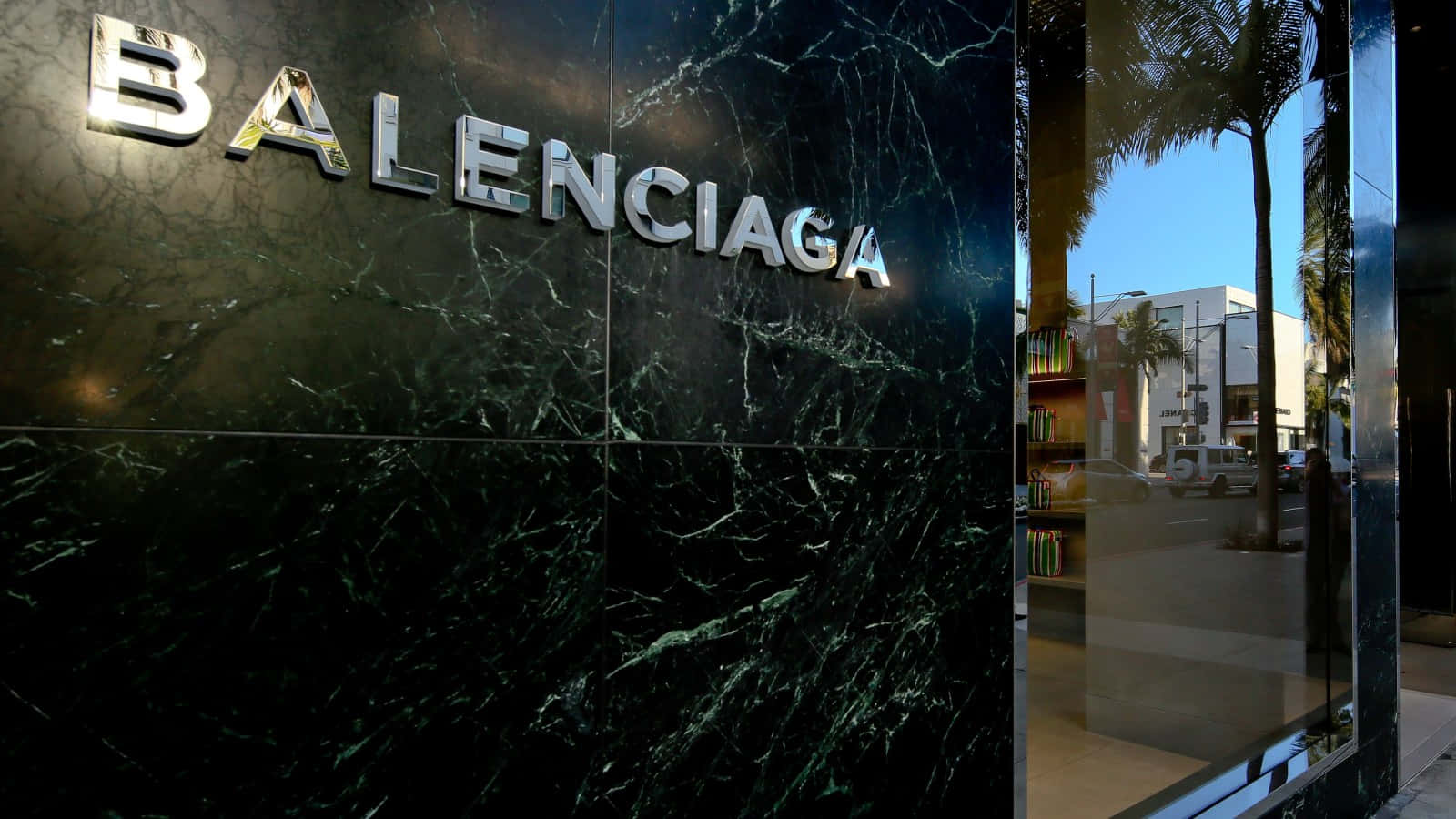 巴黎世家 桌布 壁紙 — 閃亮的鉻色 Balenciaga 標誌在深綠色大理石外牆上閃耀，旁邊是玻璃店面，映照著棕櫚樹和城市街道。