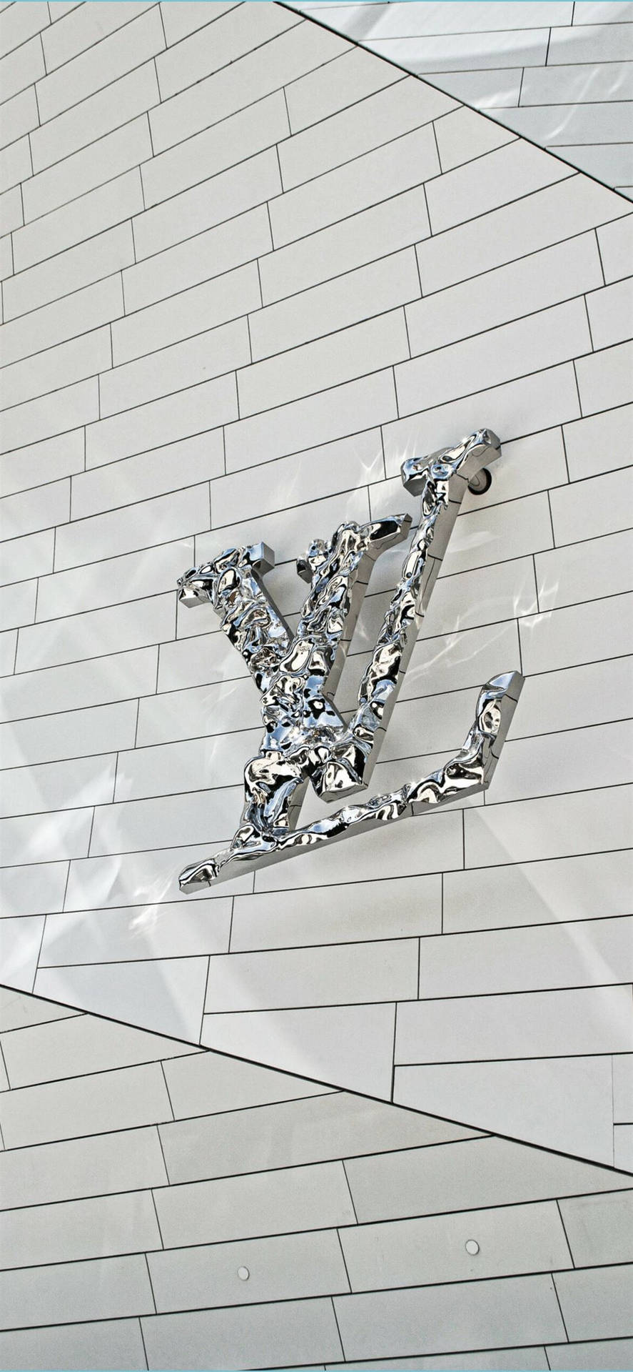  壁紙 — 閃亮的鉻色 Louis Vuitton 標誌安裝在帶有幾何對角線的白色瓷磚牆上。
