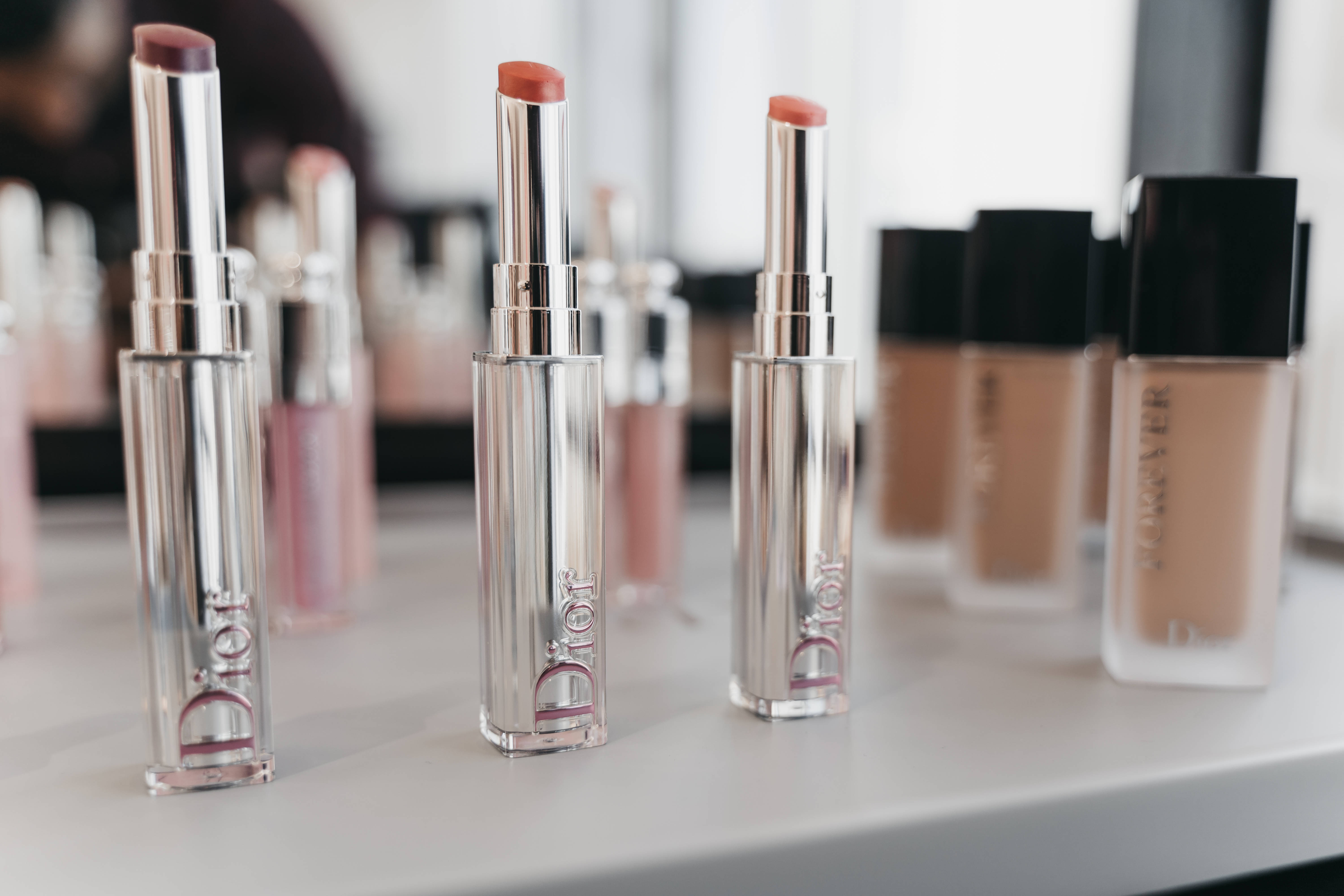 Christian Dior 桌布 壁紙 — 閃亮的銀色Dior唇膏和粉底液瓶整齊地排列在白色表面上，帶有柔和自然光。
