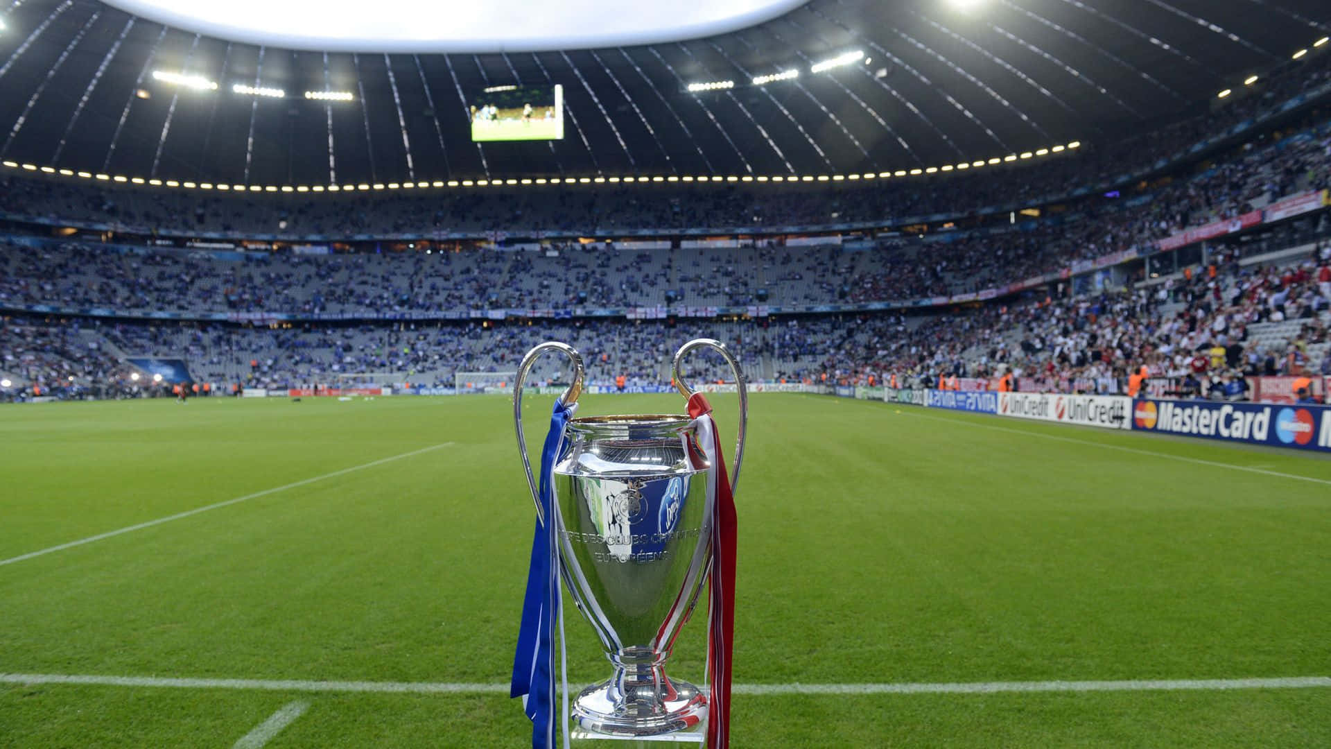 Champions League 桌布 壁紙 — 閃亮的銀色歐冠獎盃，帶有藍色和紅色絲帶，在體育場燈光下立於充滿活力的綠色足球場中央。