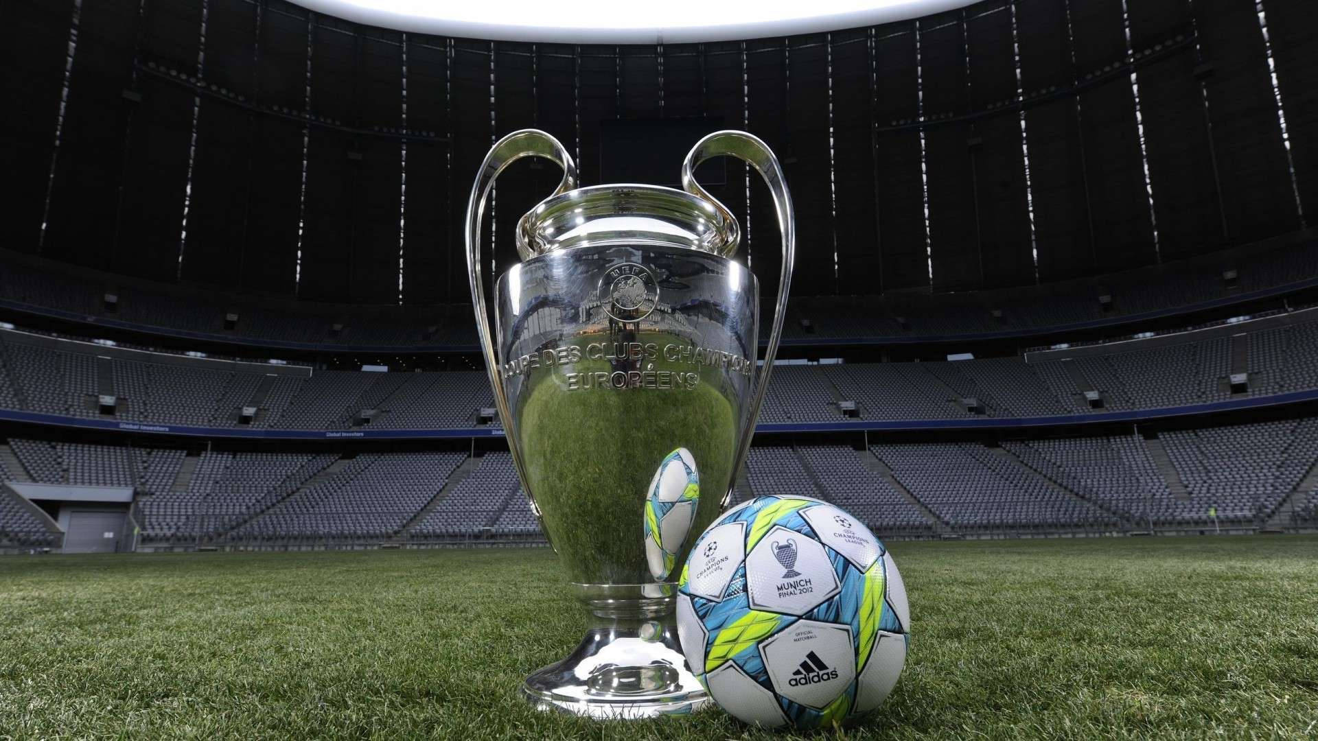 Champions League 桌布 壁紙 — 閃亮的銀色歐冠獎盃和官方比賽用球放在空蕩蕩的體育館內充滿活力的綠色球場上。