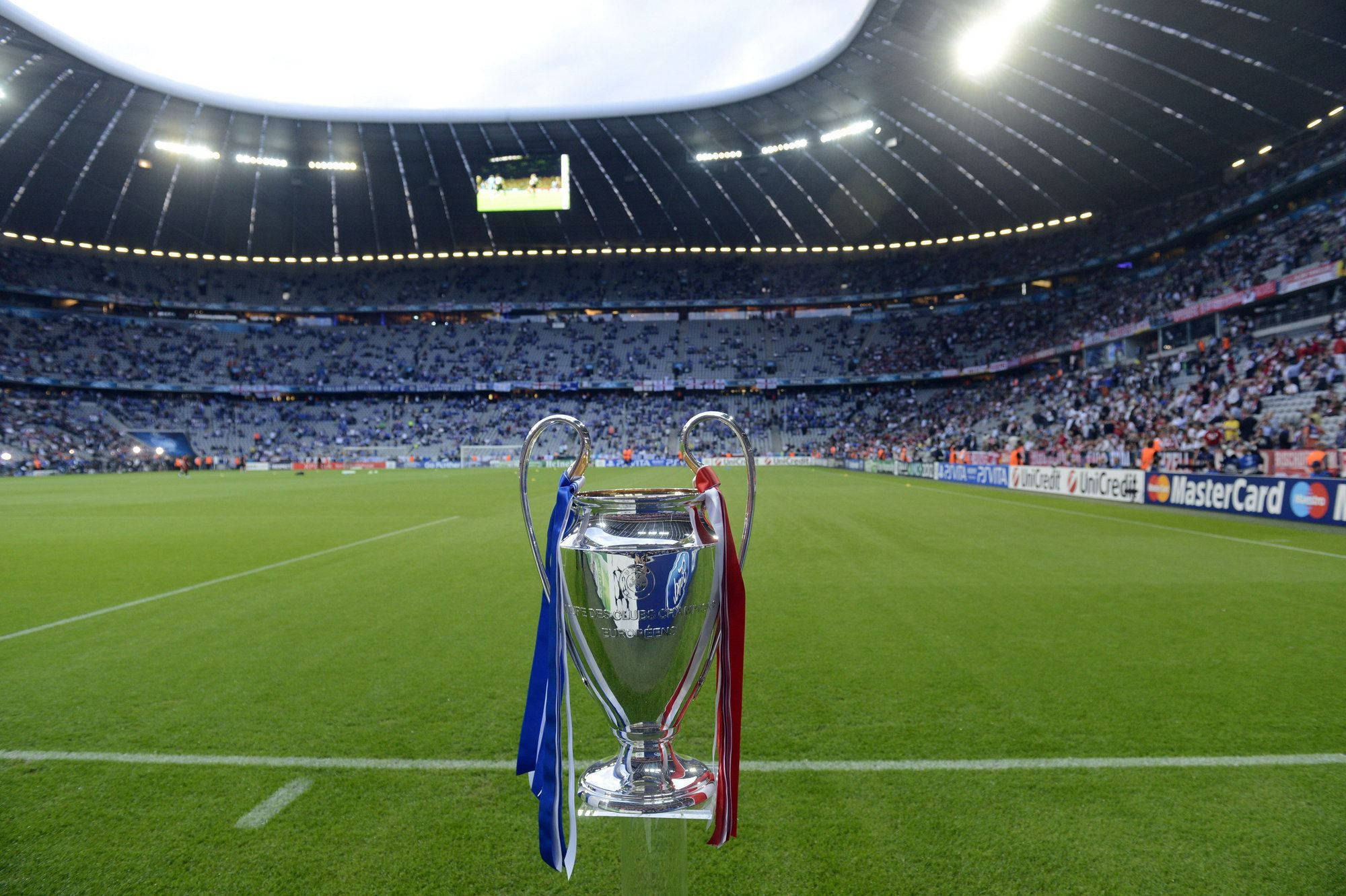 Champions League 桌布 壁紙 — 閃亮的銀色歐洲聯賽冠軍盃，帶有藍色和紅色絲帶，在體育場燈光下矗立在充滿活力的綠色足球場中央。