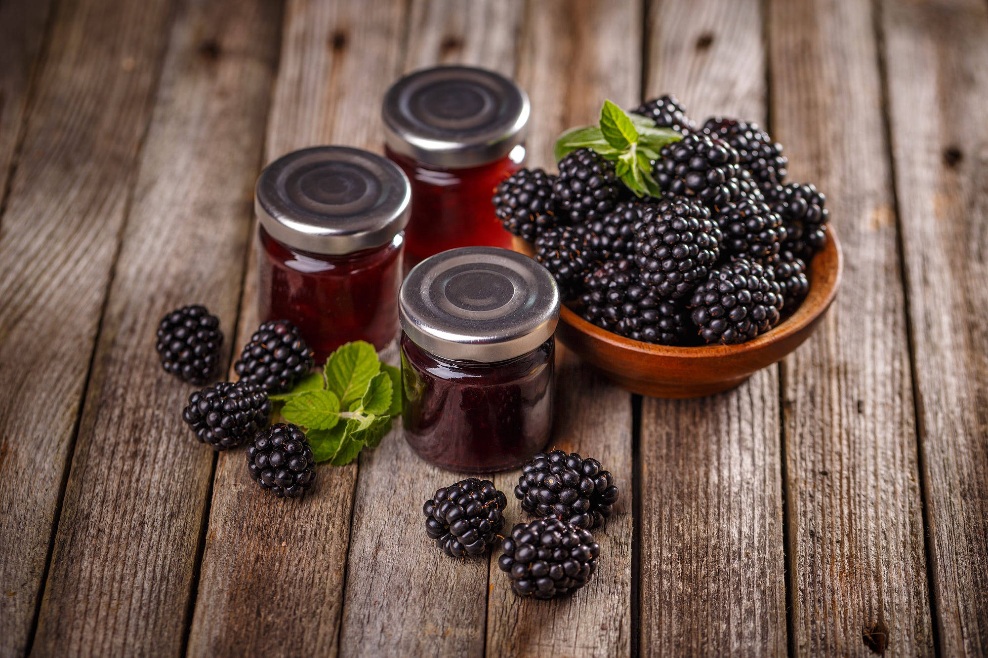 Berry 桌布 壁紙 — 閃亮的黑莓果醬罐放在一個木碗旁邊，碗裡裝滿了新鮮黑莓和薄荷，放在一張風化的木桌上。