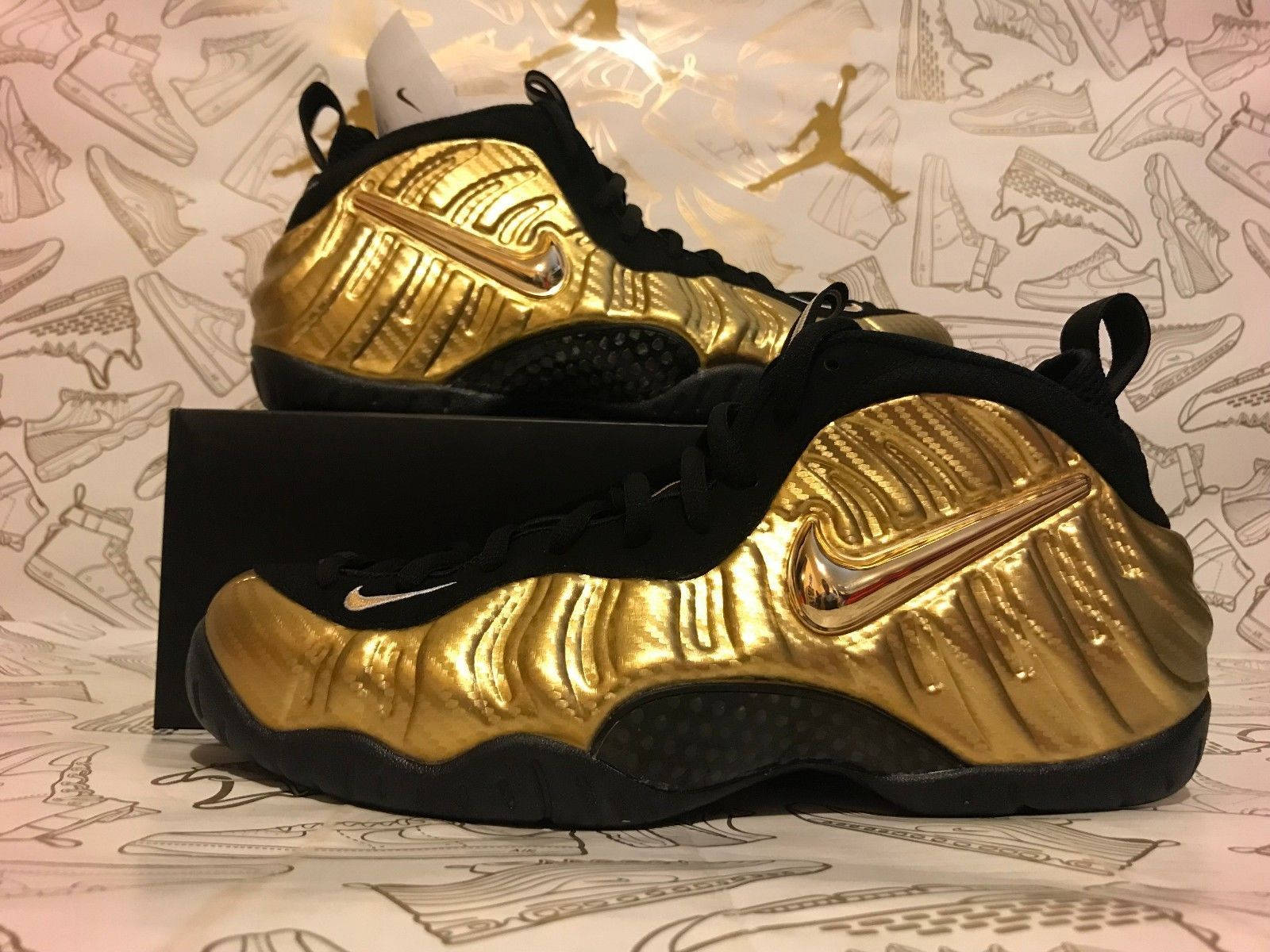 Jordan球鞋 桌布 壁紙 — 閃亮金色和黑色的Nike Air Foamposite運動鞋展示在盒子上，背景是帶有鞋類草圖圖案的壁紙。
