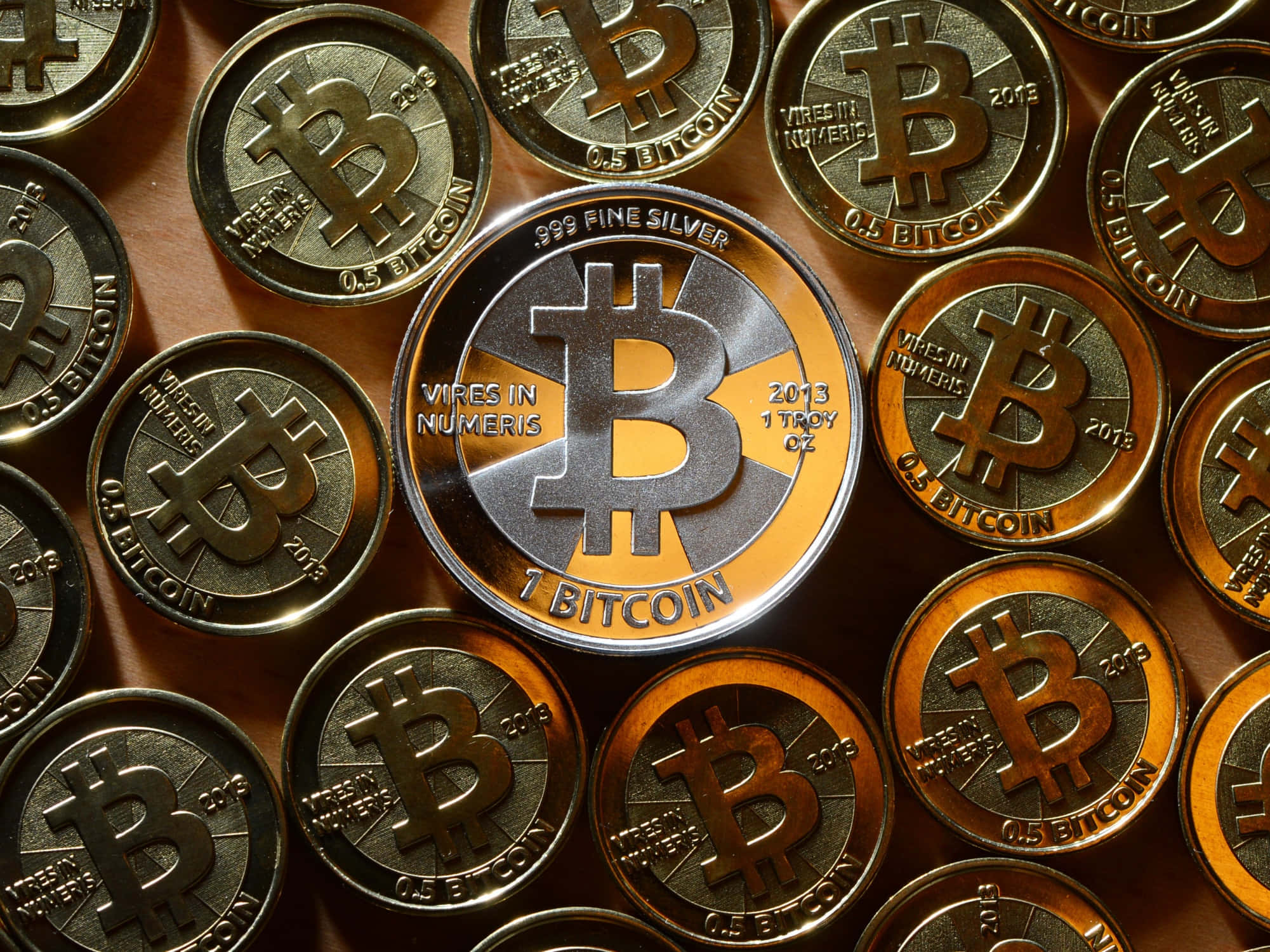 比特幣 桌布 壁紙 — 閃亮銀色和金色比特幣硬幣的特寫，上面刻有「1 BITCOIN」和「0.5 BITCOIN」字樣，以密集圖案排列。