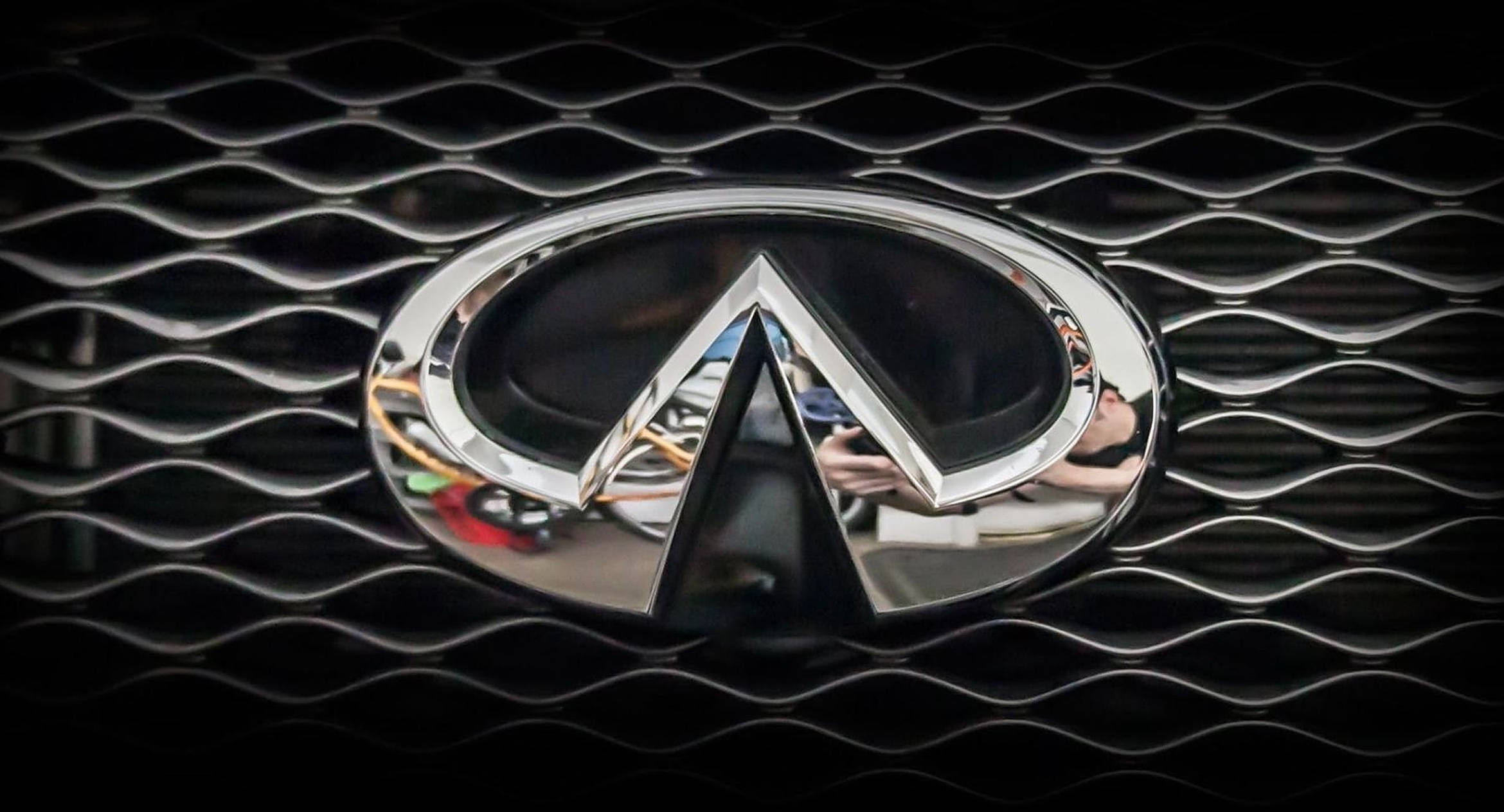 Infiniti 桌布 壁紙 — 閃亮鍍鉻Infiniti標誌居中於黑色波浪形水箱護罩上，反射表面顯示模糊的周圍環境。